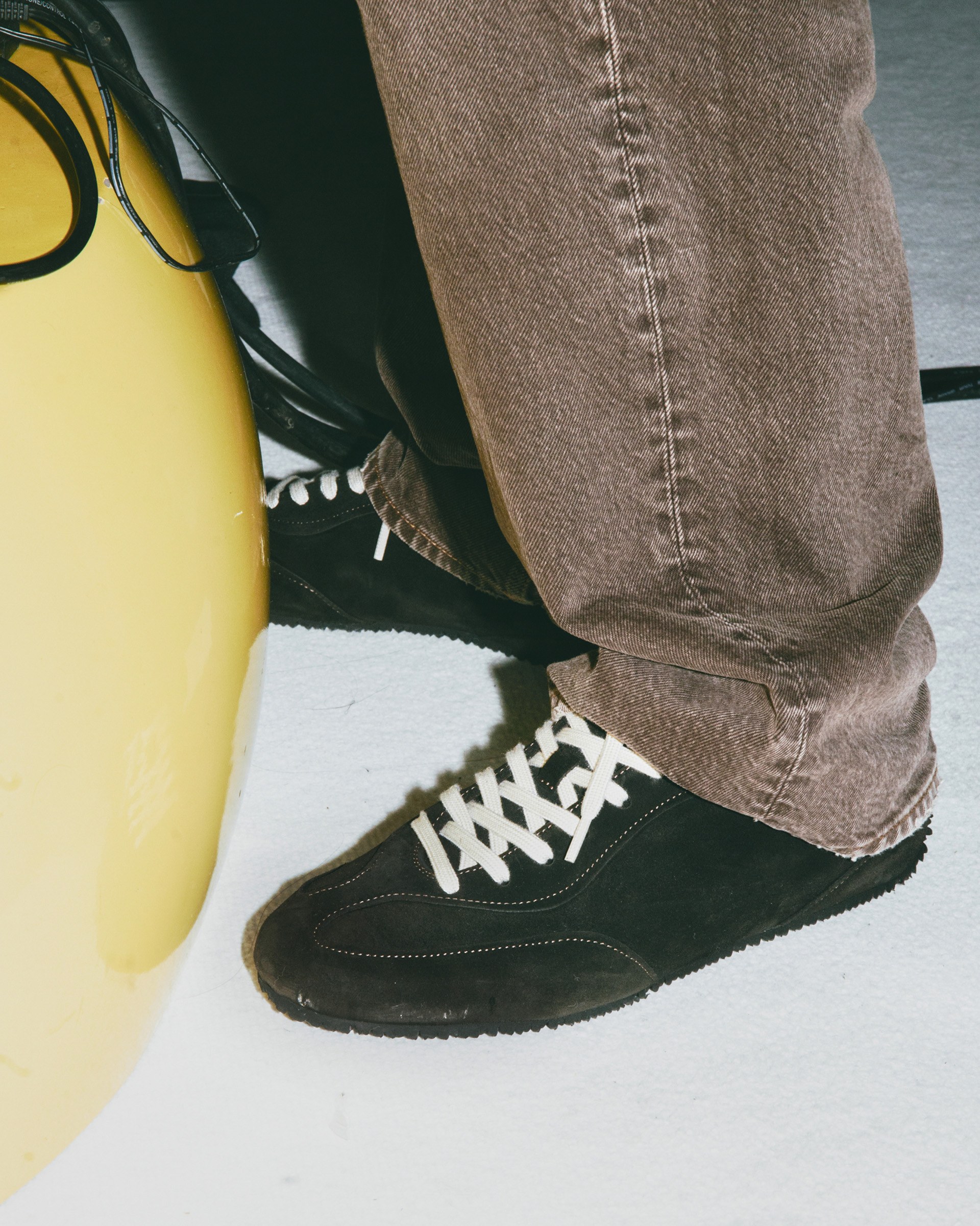 Brown suede sneakers with white laces worn by a person in brown pants, standing next to a yellow object and a black bag.