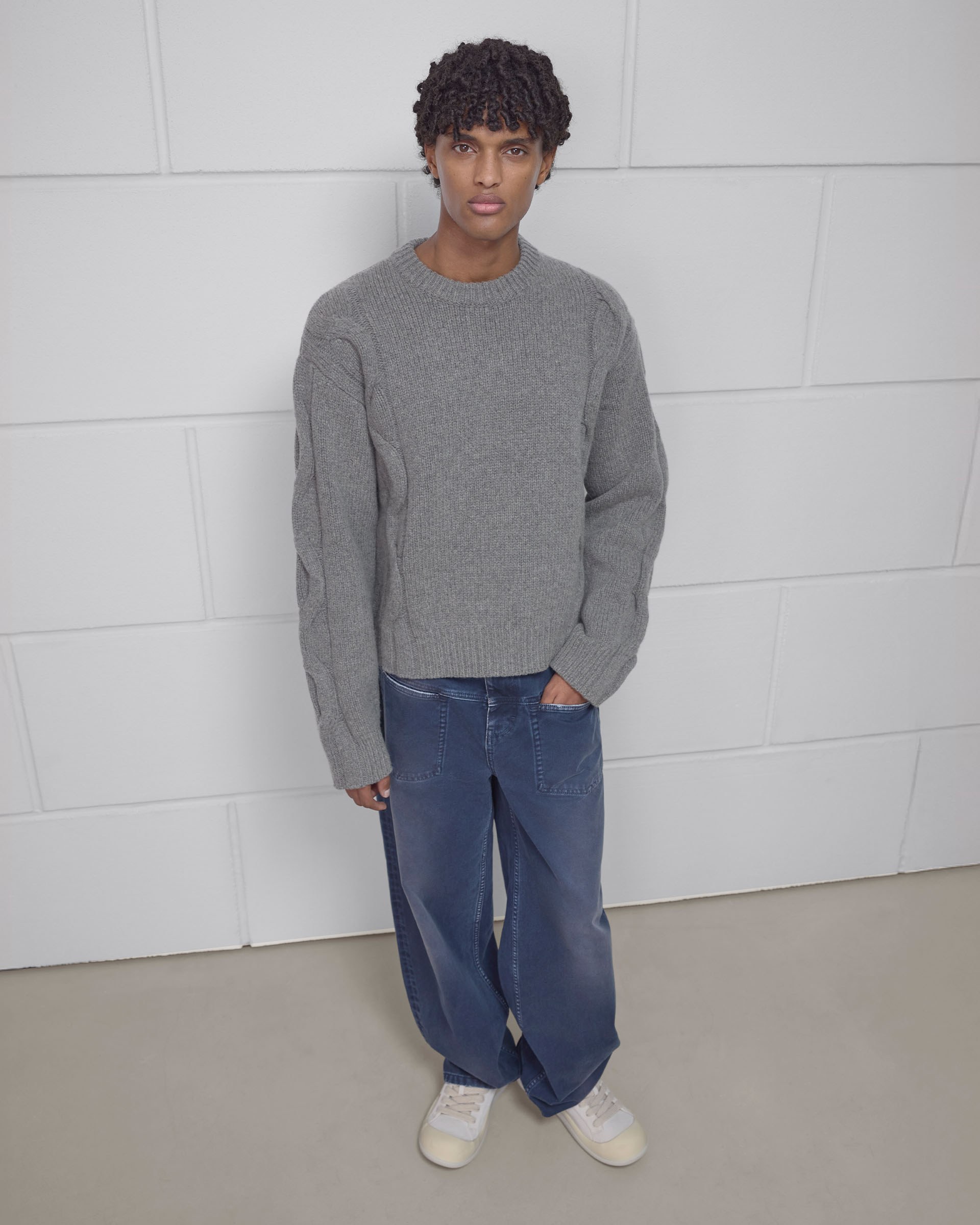 Person in a gray sweater and blue jeans stands against a white tiled wall, with hands in pockets.