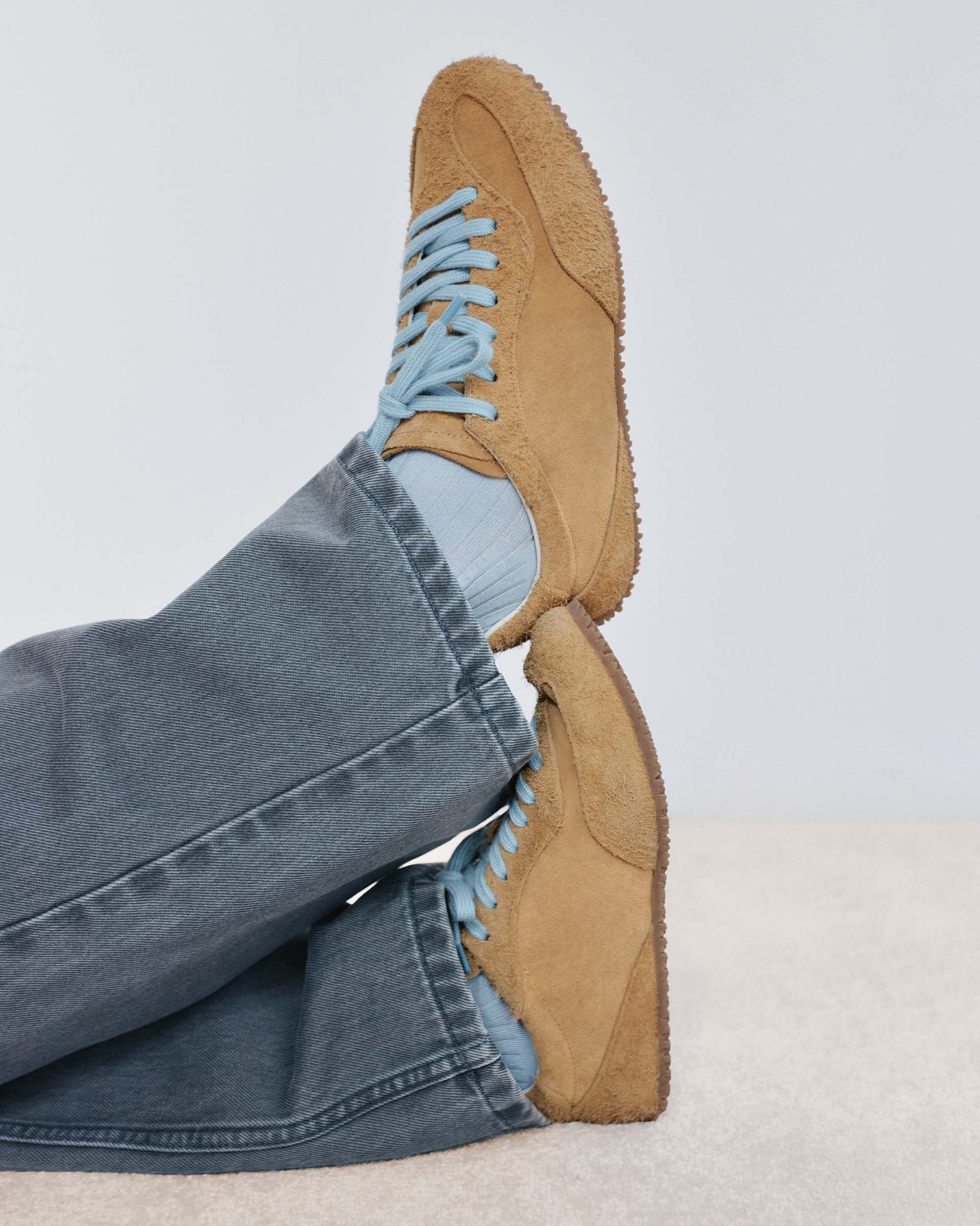 Person wearing beige suede sneakers with blue laces, light blue socks, and gray jeans, legs crossed on a light surface.