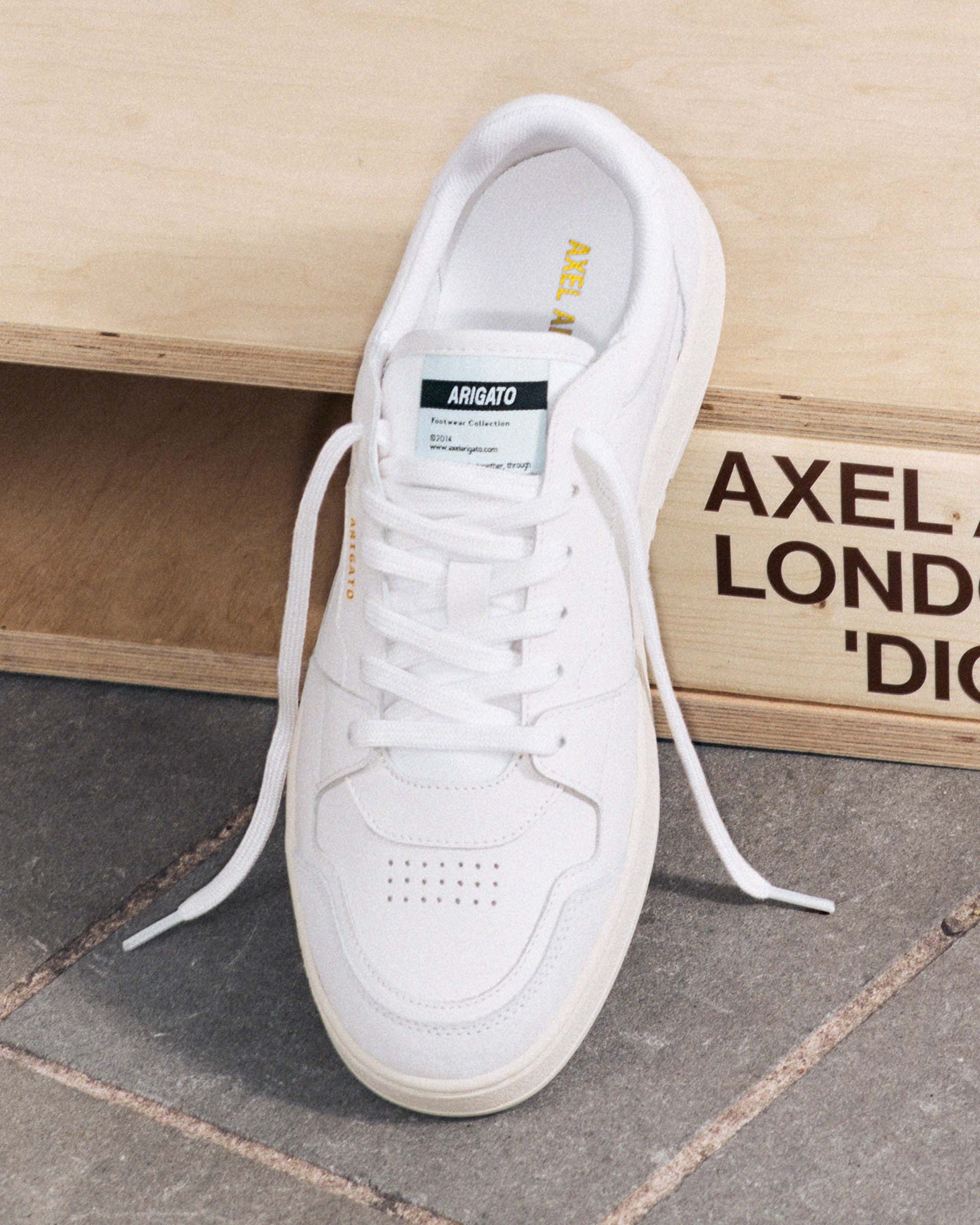 White sneaker with laces on a stone surface, featuring a green label on the tongue and a wooden crate in the background.