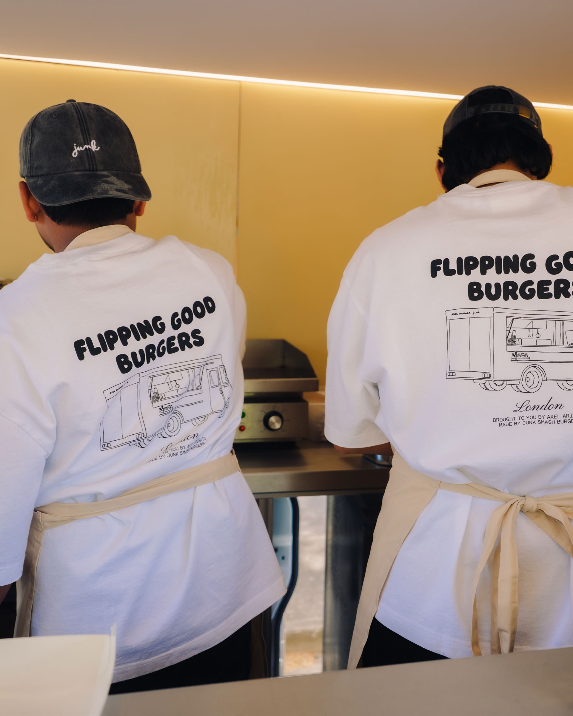 Two people wear "Flipping Good Burgers" shirts and aprons, working at a grill in a food truck.