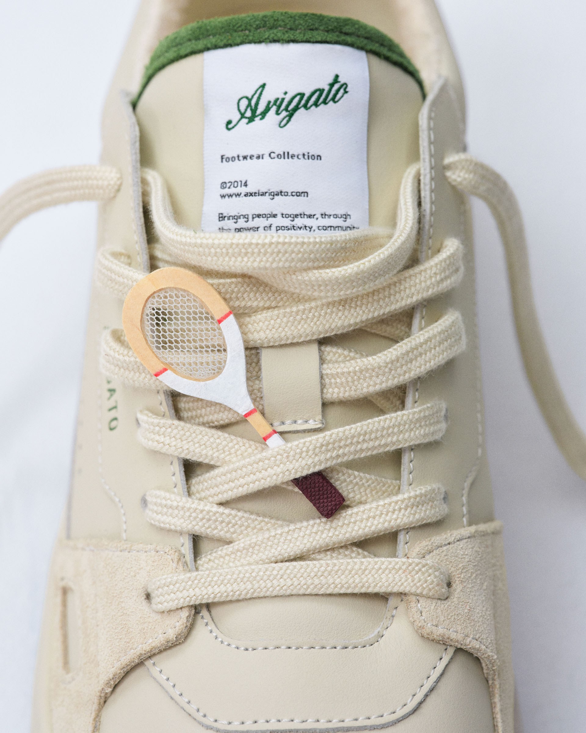 Close-up of a beige sneaker with the brand "Arigato" on the tongue and a miniature tennis racket charm woven into the laces.