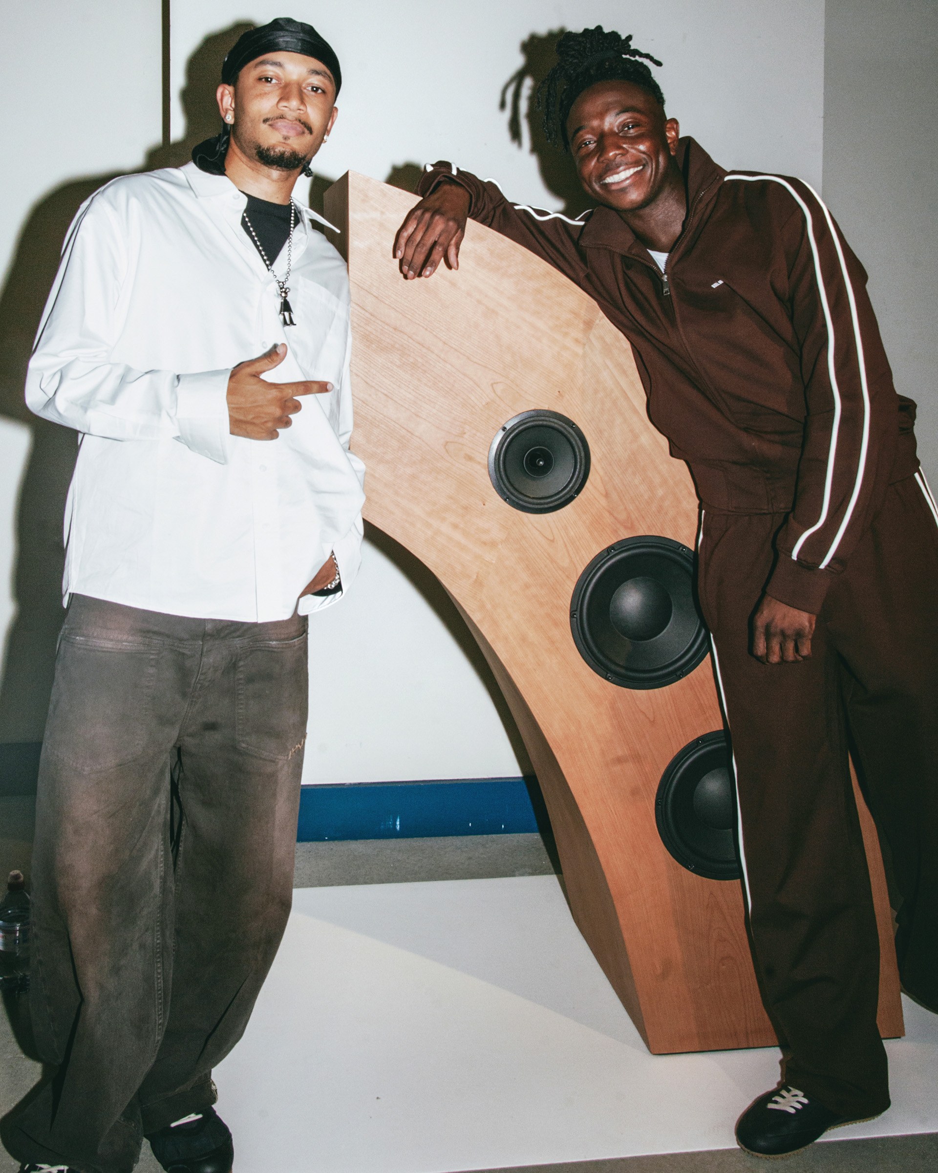Two men pose with a uniquely designed wooden speaker, smiling and casually dressed, one in a tracksuit and the other in a white shirt.
