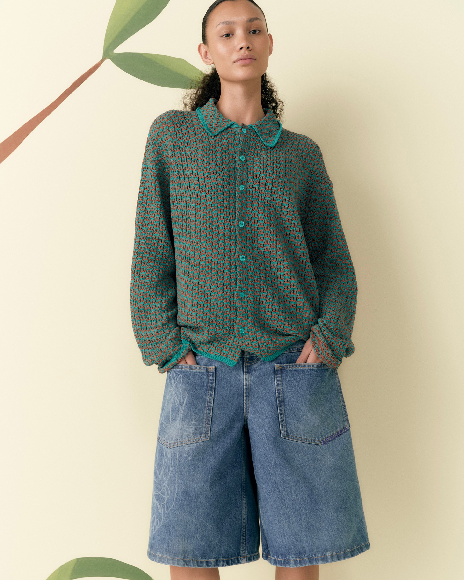 A person wearing a green patterned sweater and denim knee-length shorts stands against a light-colored background with leaf designs.