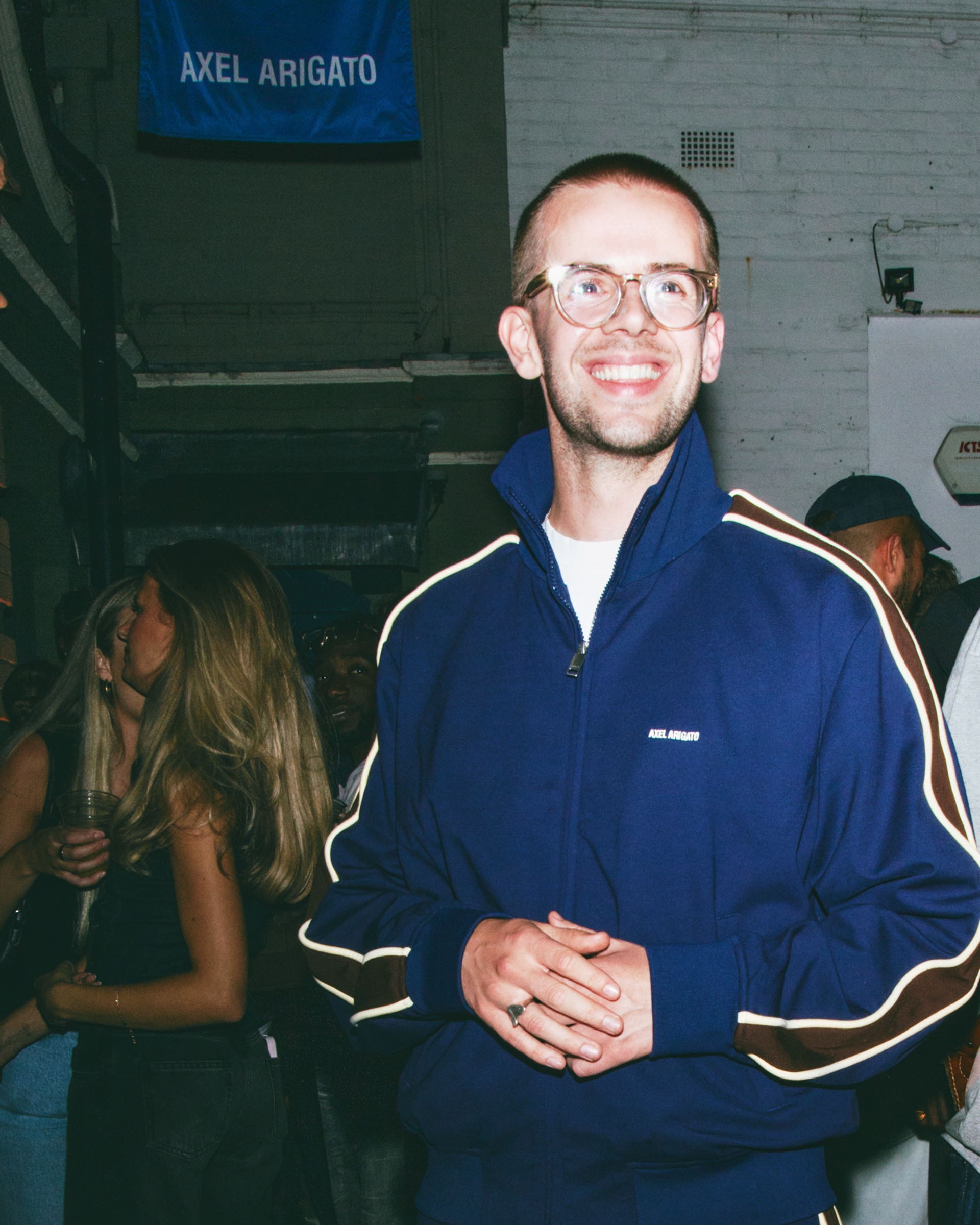 A person wearing glasses and a blue track jacket smiles at an indoor event, with people and an "Axel Arigato" banner in the background.
