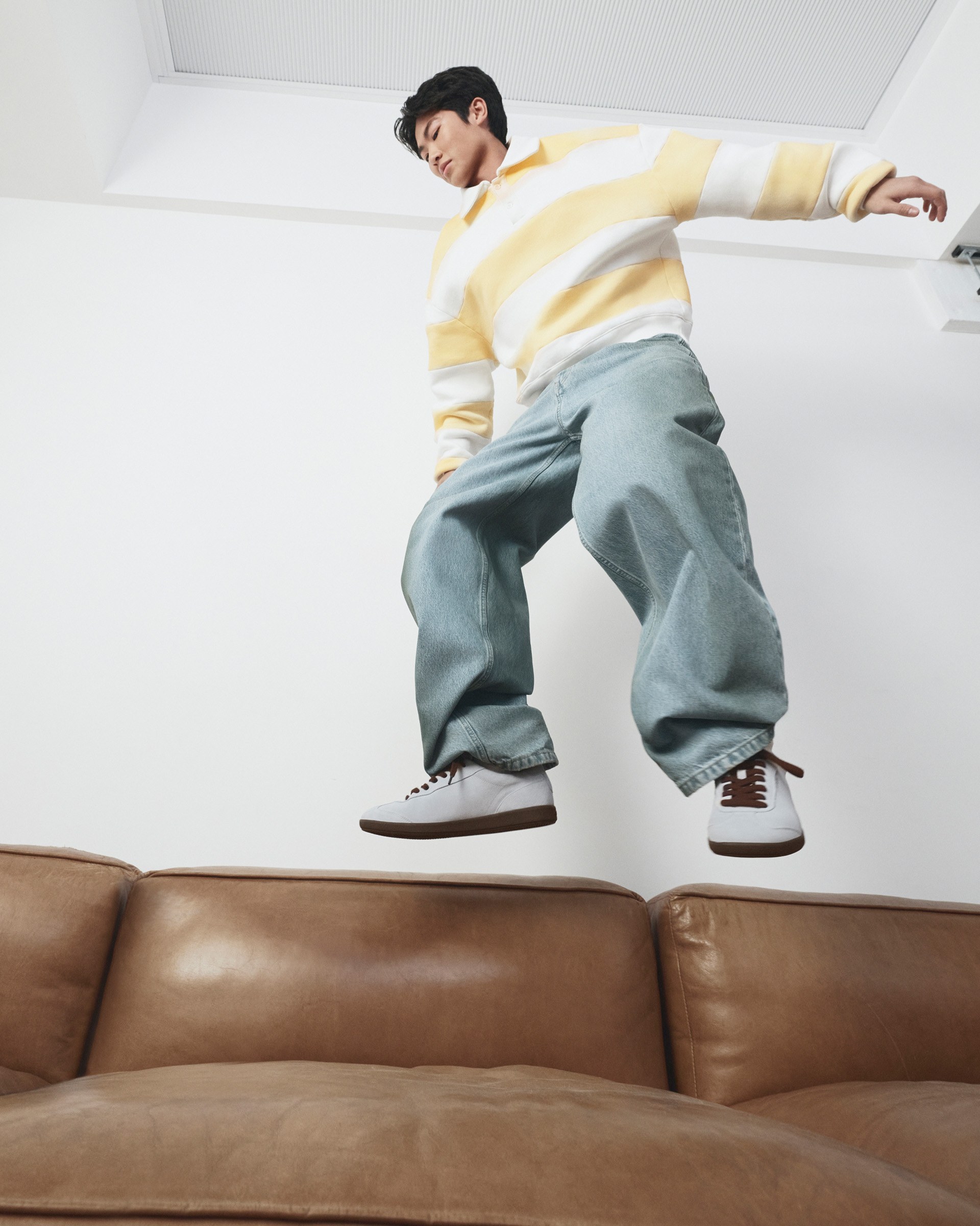 Person in a yellow and white sweater and baggy jeans mid-jump over a brown leather couch in a white room.