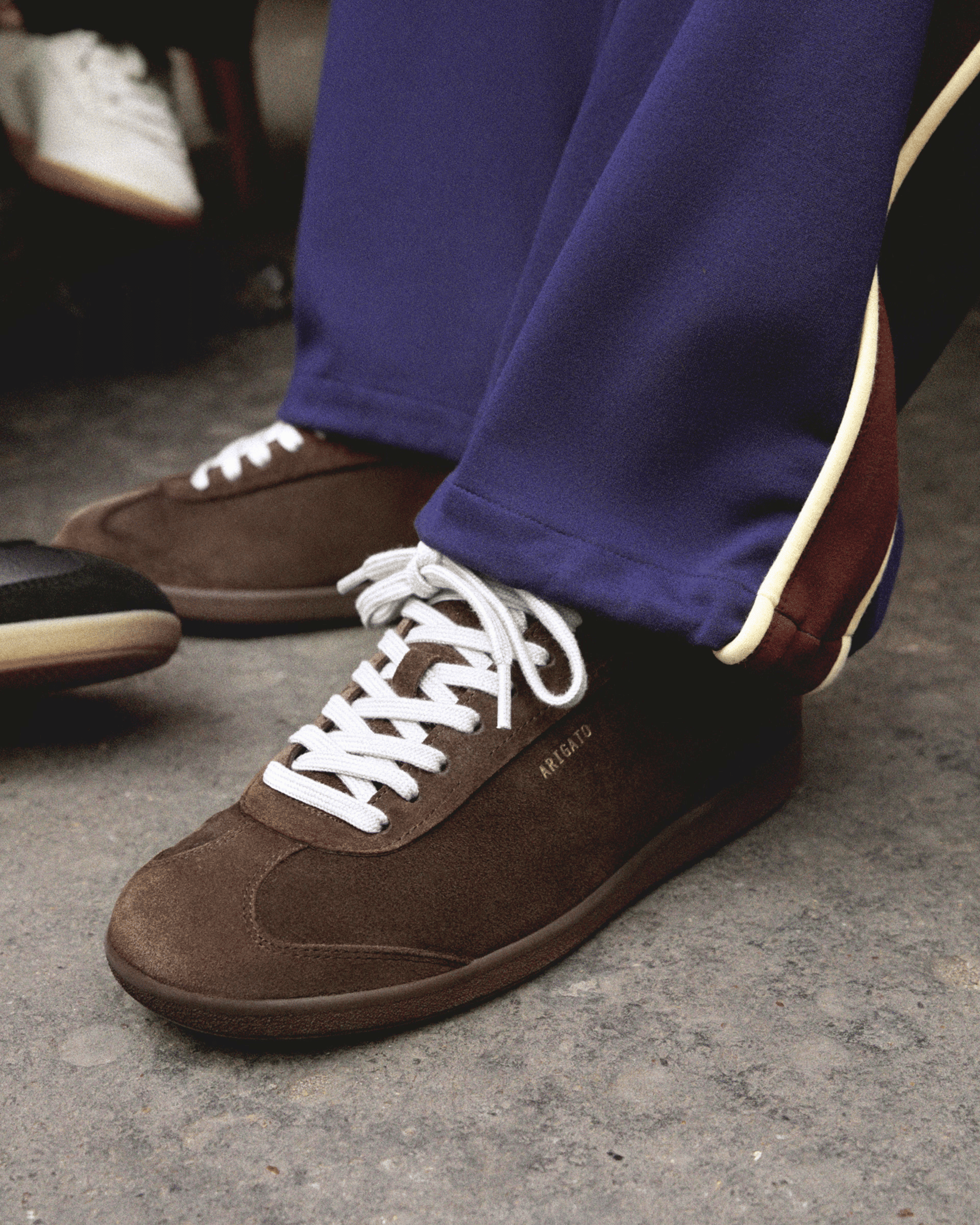 Brown sneakers with white laces, worn with blue pants with white trim, on a textured gray floor.