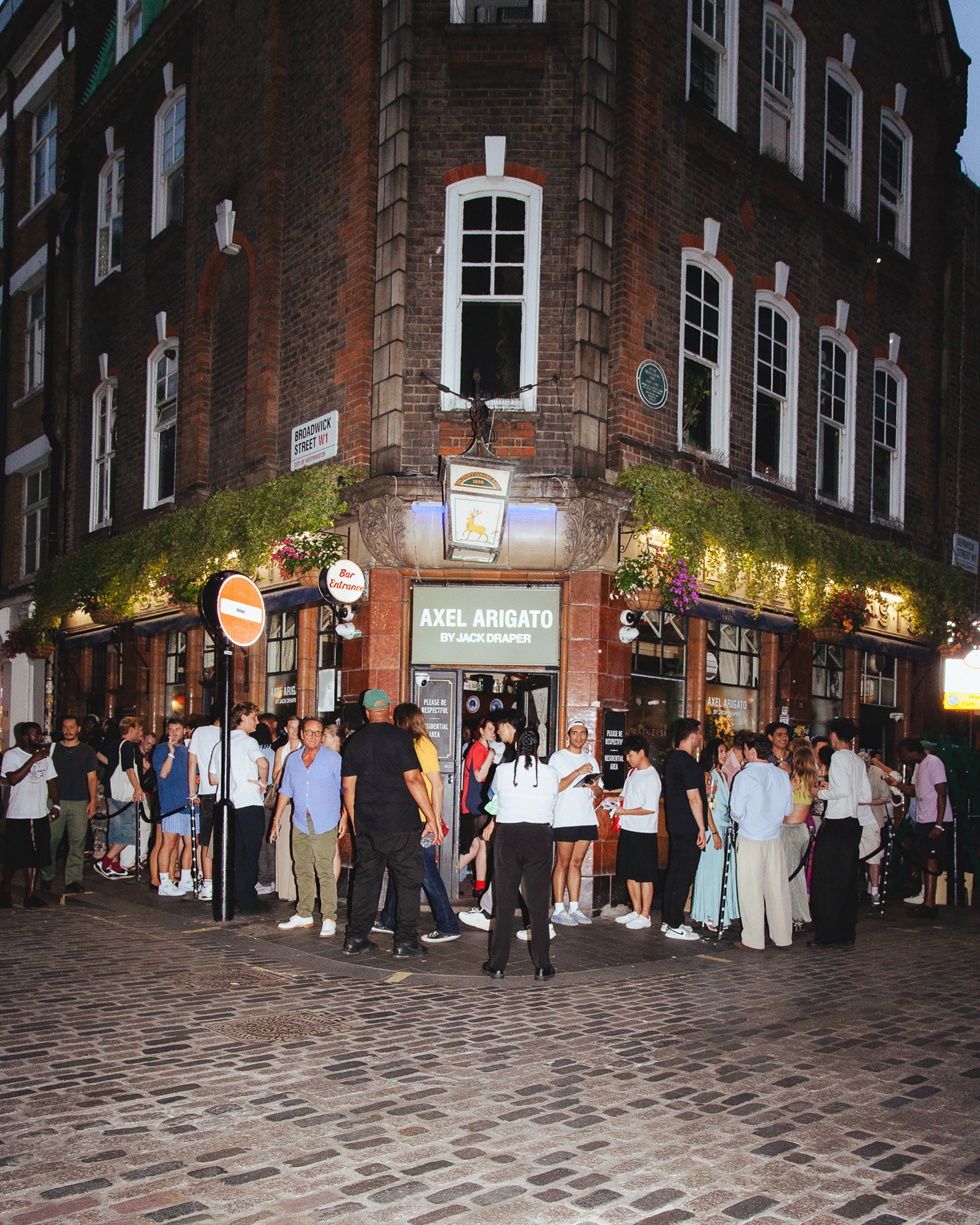 A lively crowd gathers outside a shop on a cobblestone corner in the evening, with greenery and signage above the entrance.