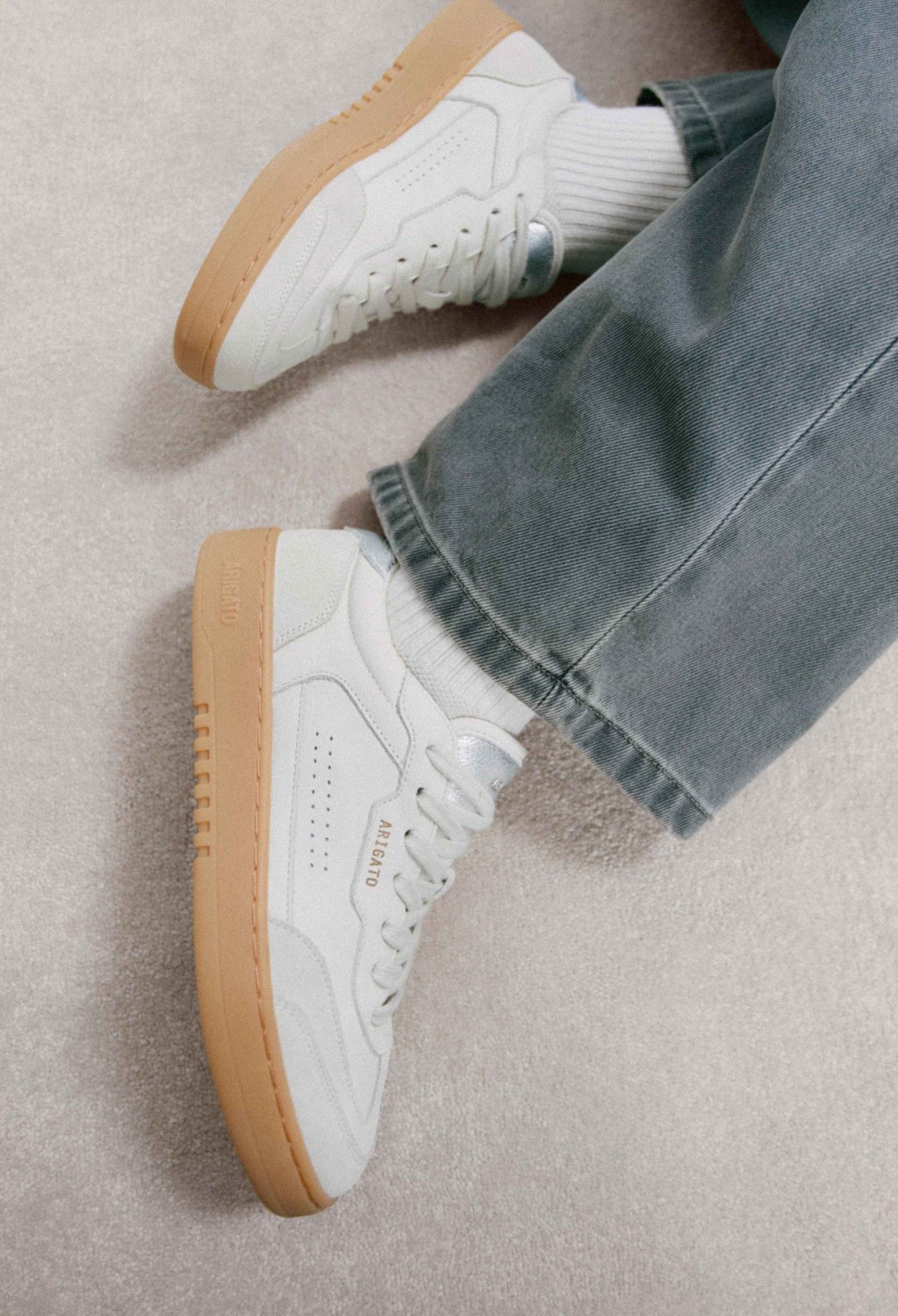 White sneakers with gum soles worn with light gray jeans and white socks on a beige carpet.