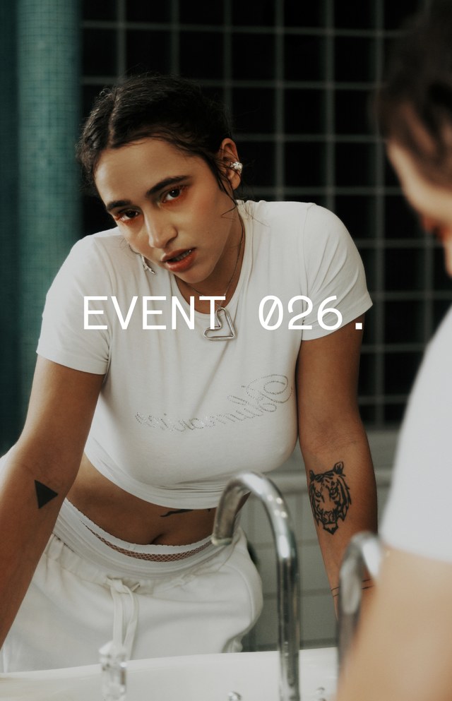 Woman in a white t-shirt leans over a sink with "EVENT 026" text overlay, set in a tiled bathroom background.