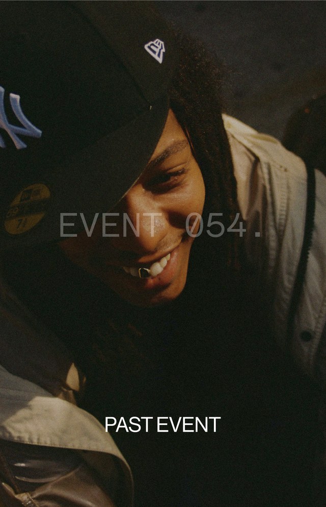 Person smiling, wearing a black cap with a logo, and a jacket. Text overlay reads "EVENT 054" and "PAST EVENT." Dimly lit setting.