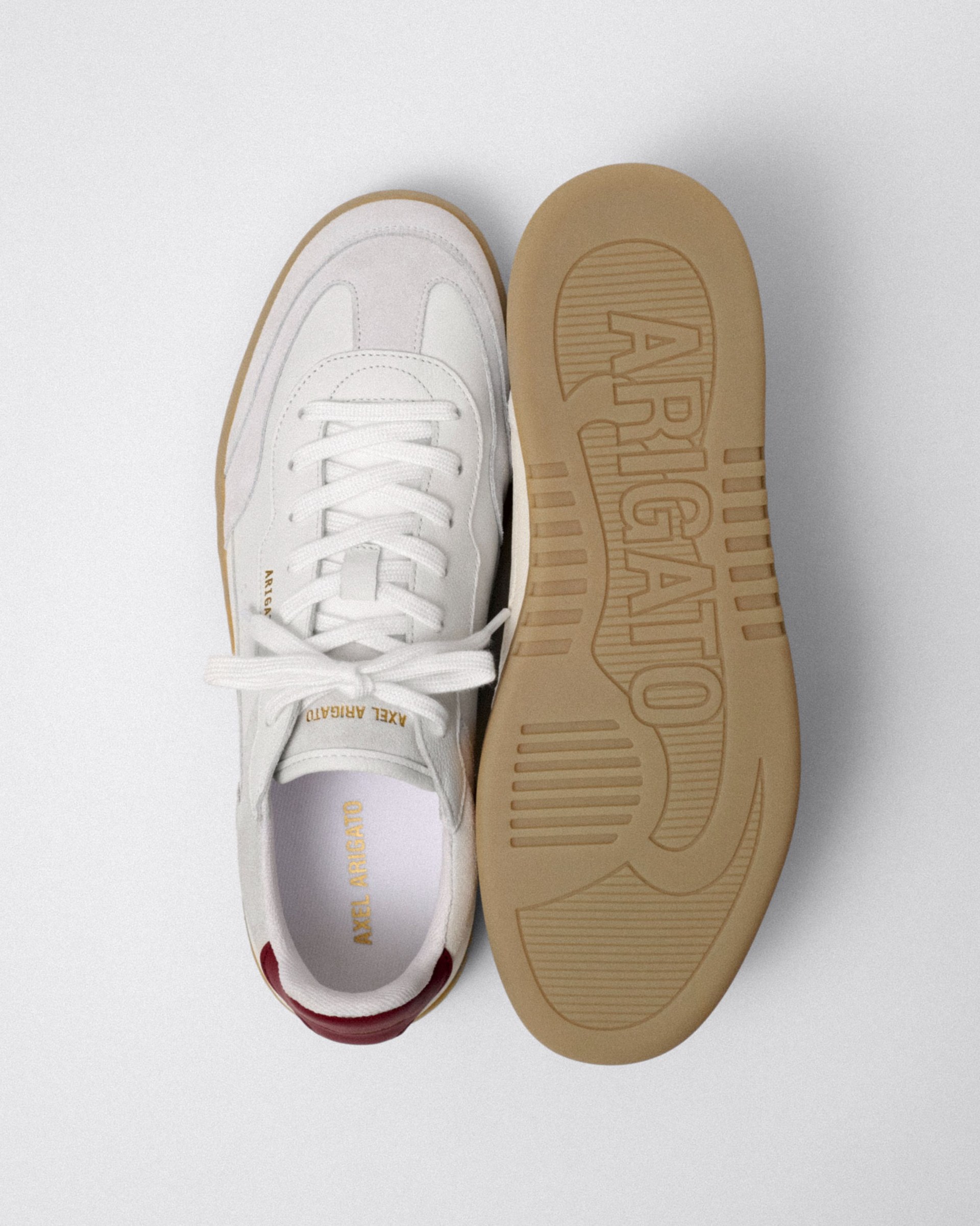White sneakers with gum sole, displayed from above, one showing the top and the other its sole imprinted with "Arigato", red heel detail.
