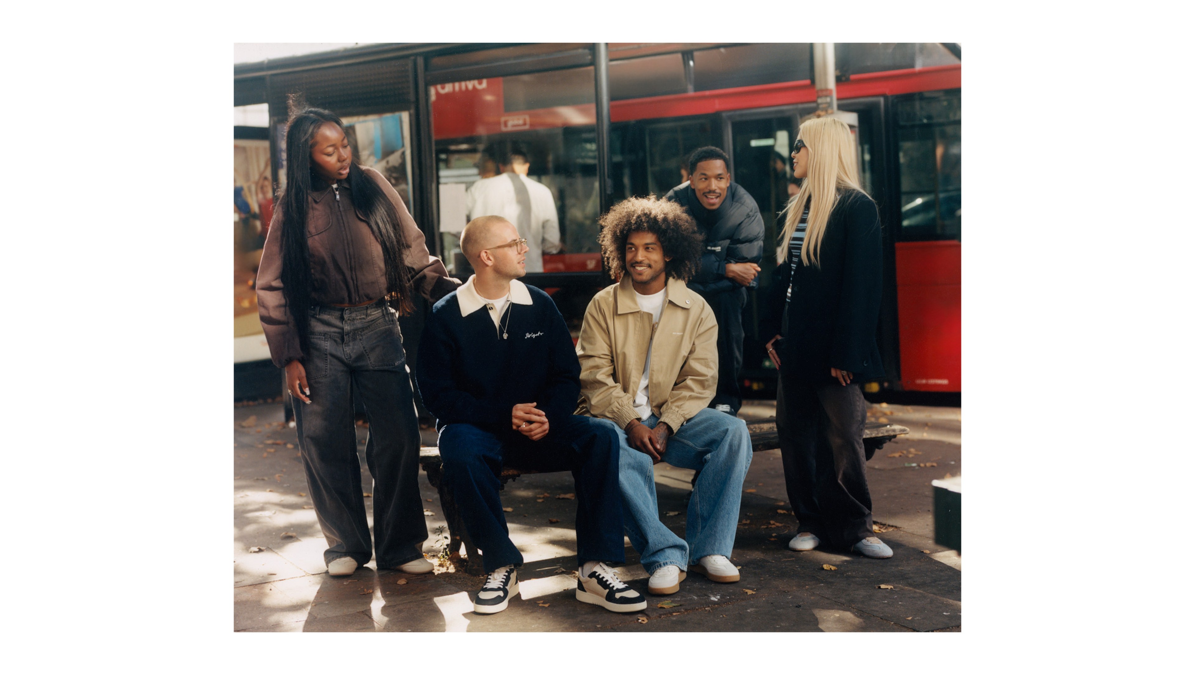 A group of people socializing near a bus stop. Two sit on a bench while three stand, all dressed in casual, stylish clothing.
