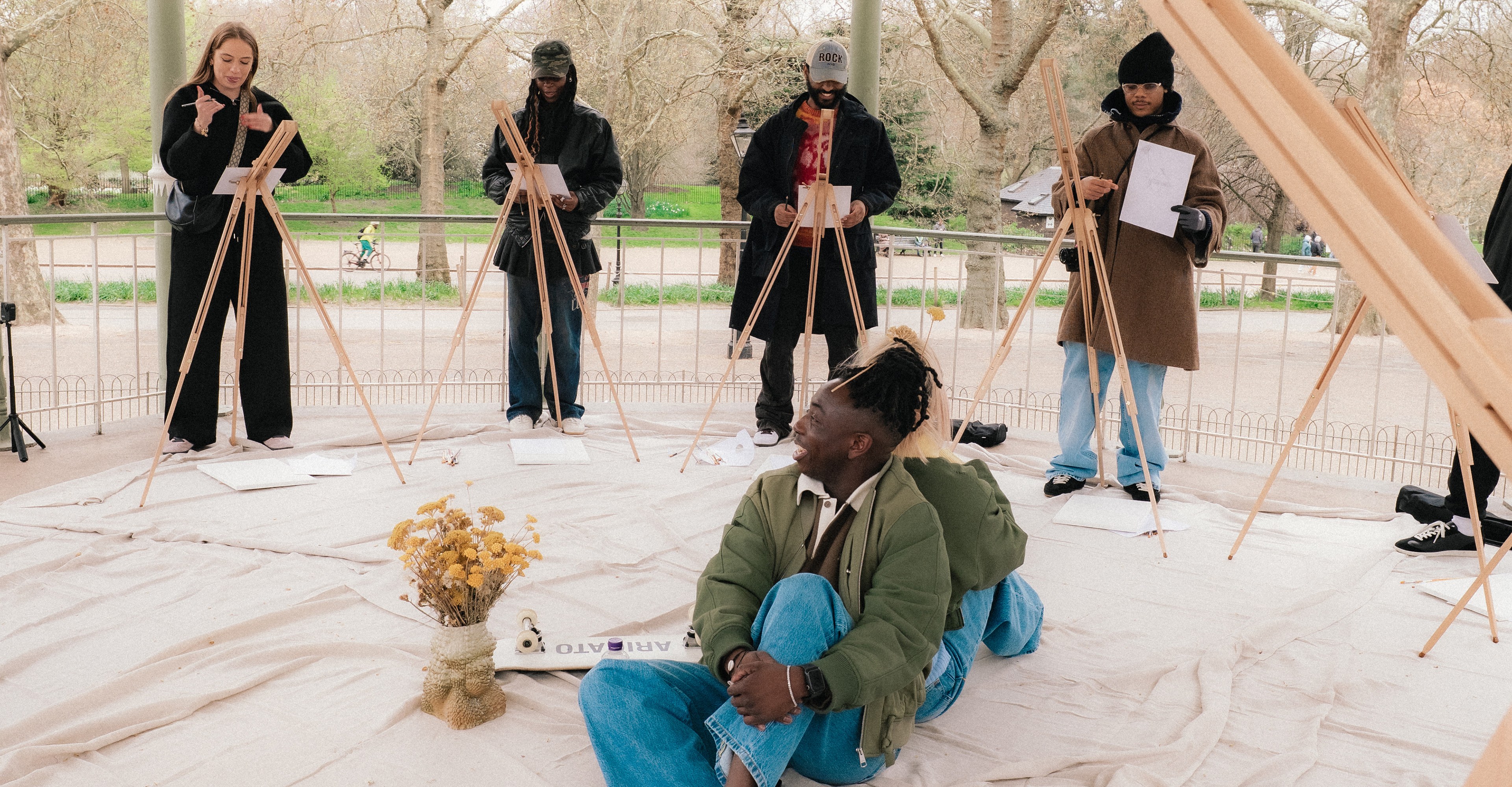 A group of people sketch on easels in a park gazebo, with one person seated in the center on a sheet, surrounded by art supplies and dried flowers.