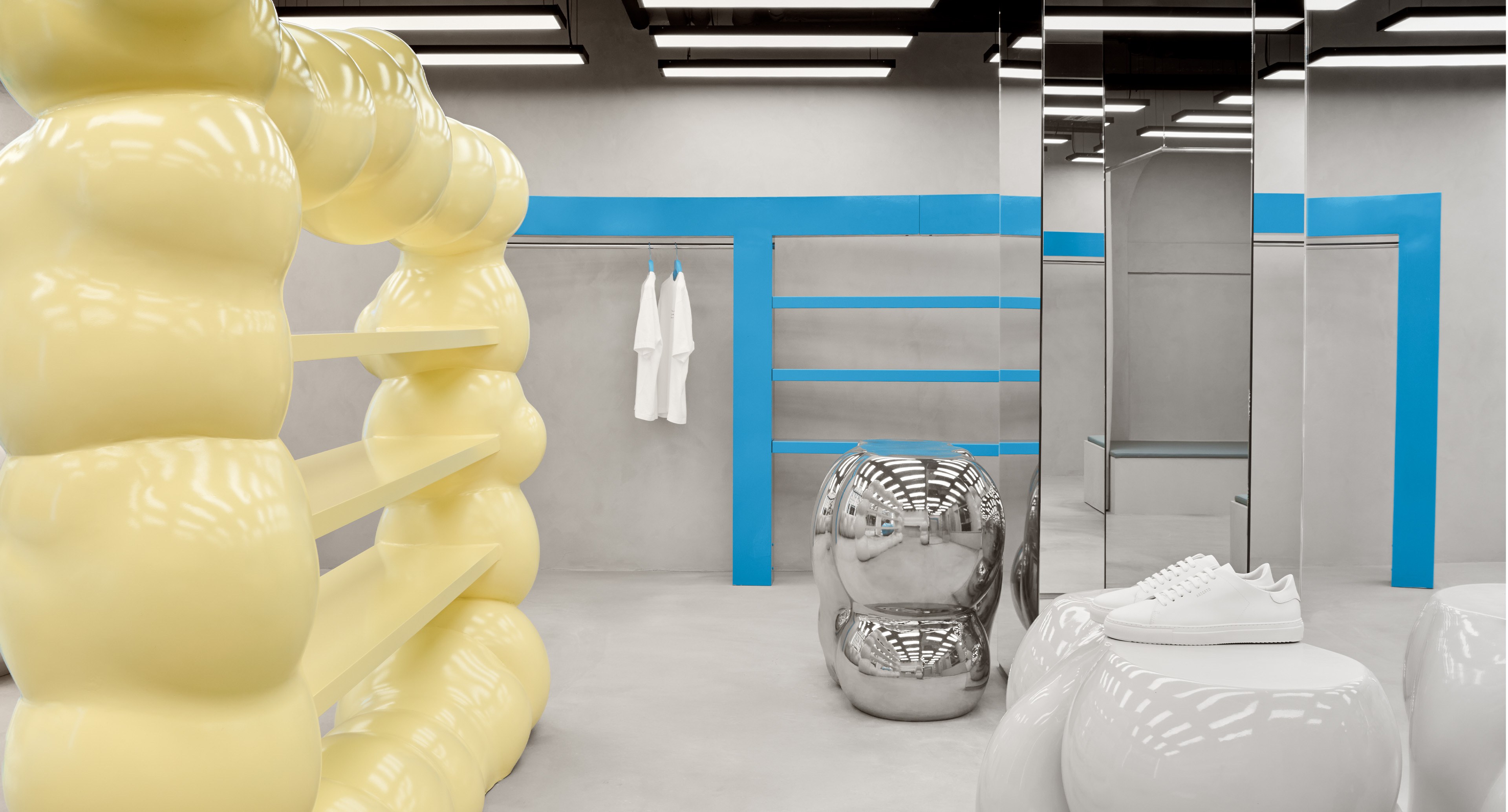 Modern store interior with unique yellow shelving, a blue-framed display, metallic sculptures, and a pair of white sneakers on a pedestal.