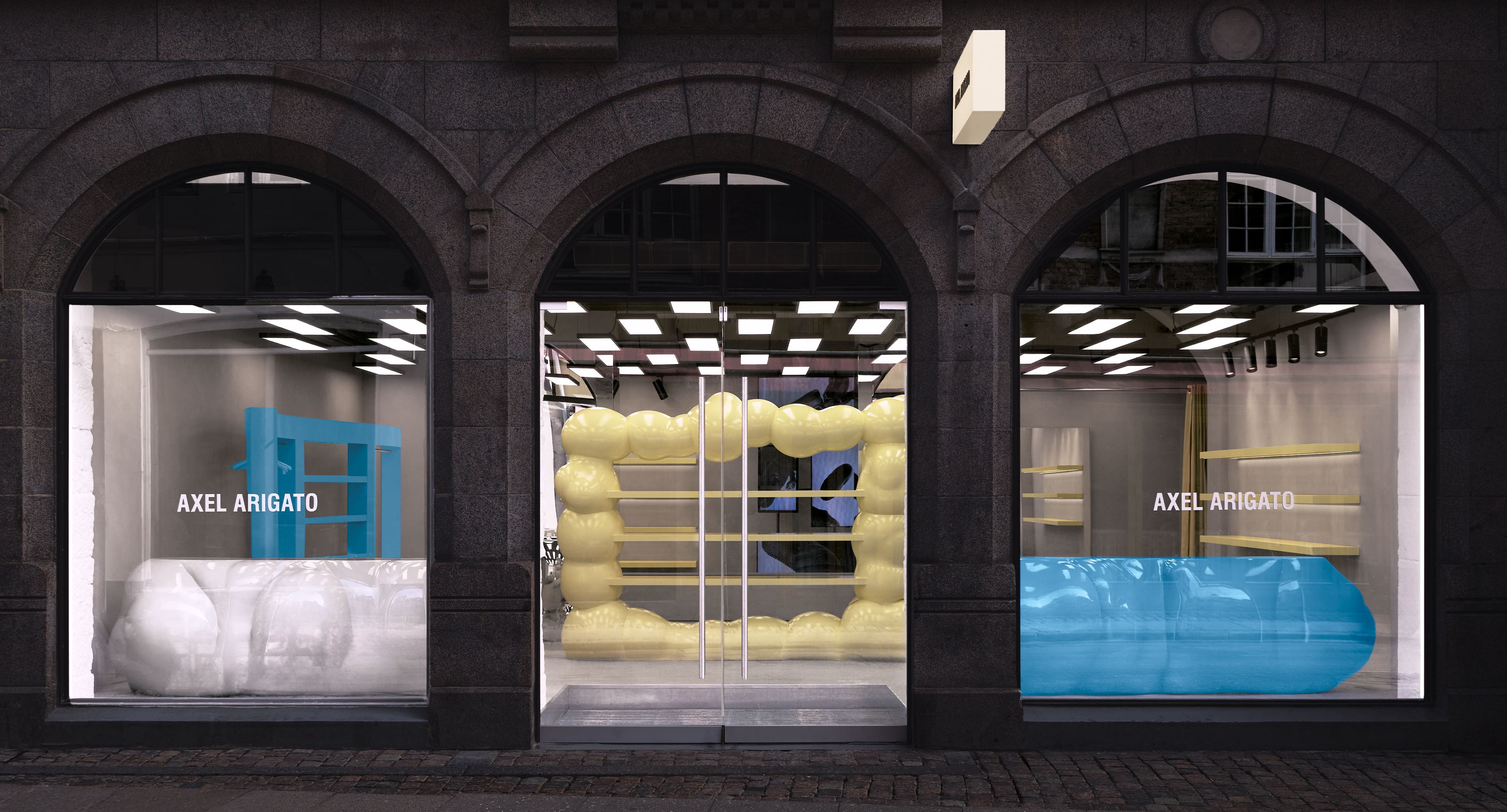 Storefront with three arched windows displaying modern installations in blue, yellow, and white, labeled "Axel Arigato."