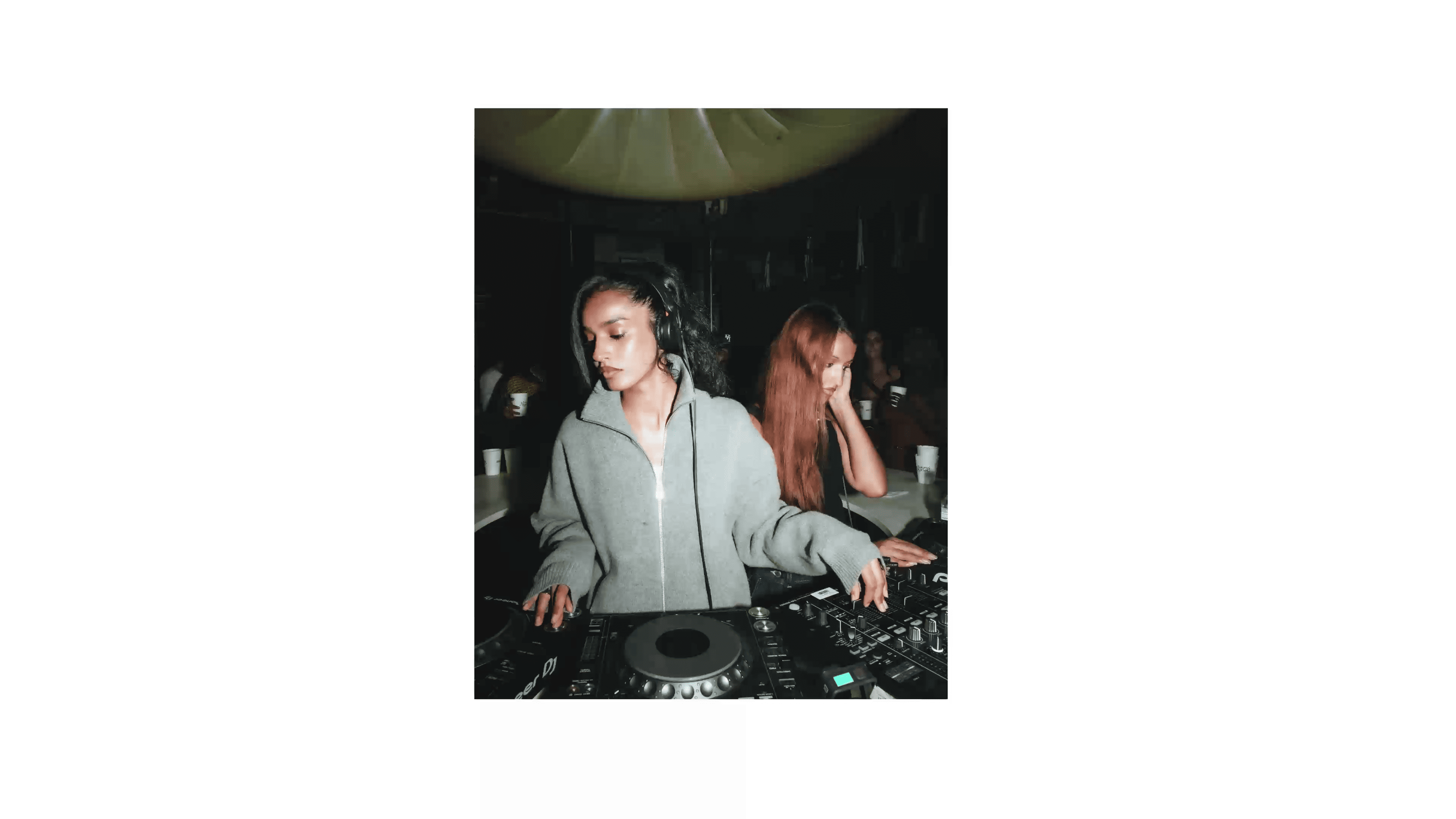 Two DJs are performing at a dimly lit venue, focused on mixing music, surrounded by equipment.