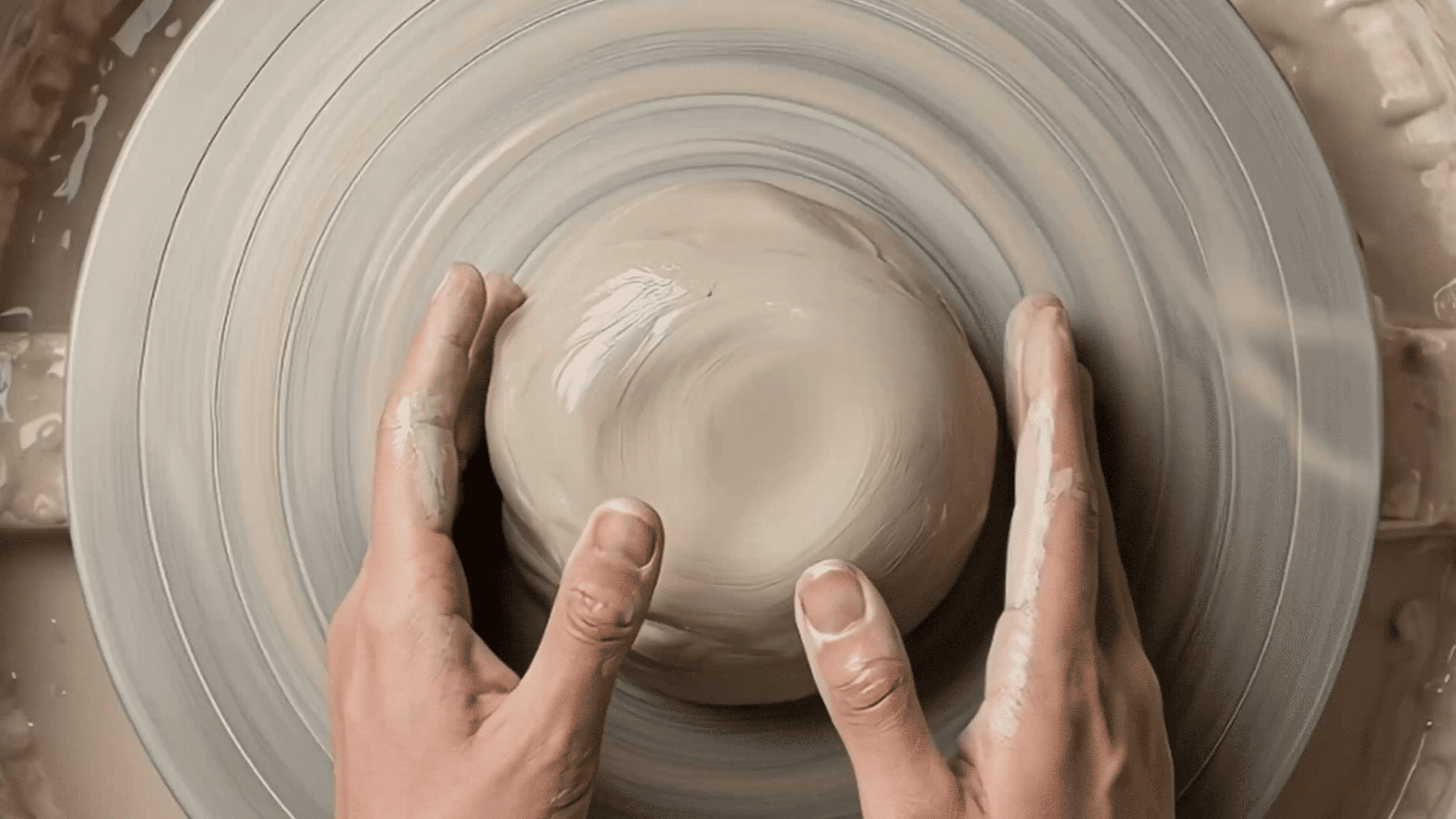 Hands shaping wet clay on a spinning pottery wheel, with a smooth, circular motion visible, creating a bowl-like shape.