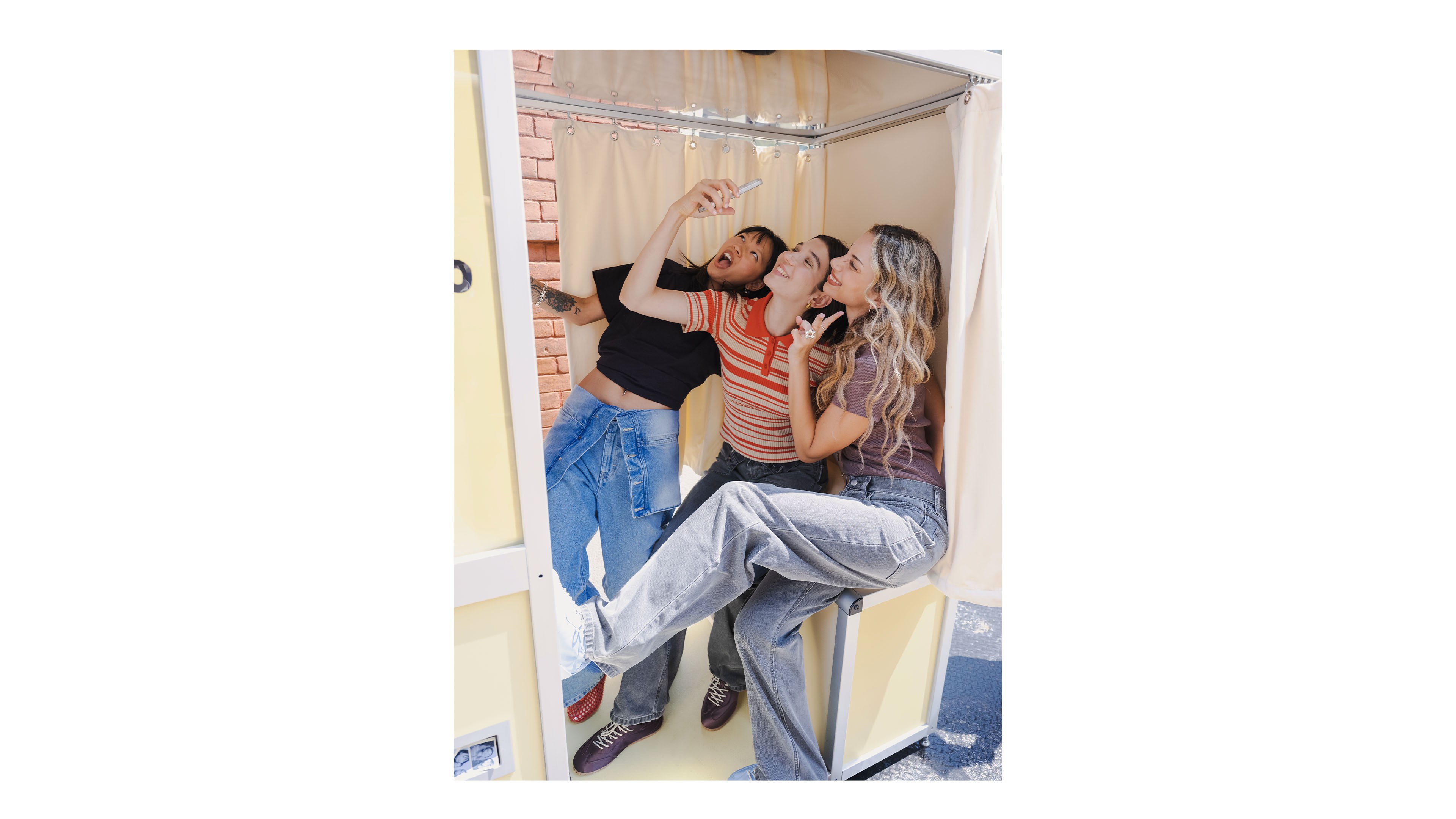 Three friends pose playfully inside a photo booth, taking a selfie. They are smiling and dressed casually, with one person lifting their foot.