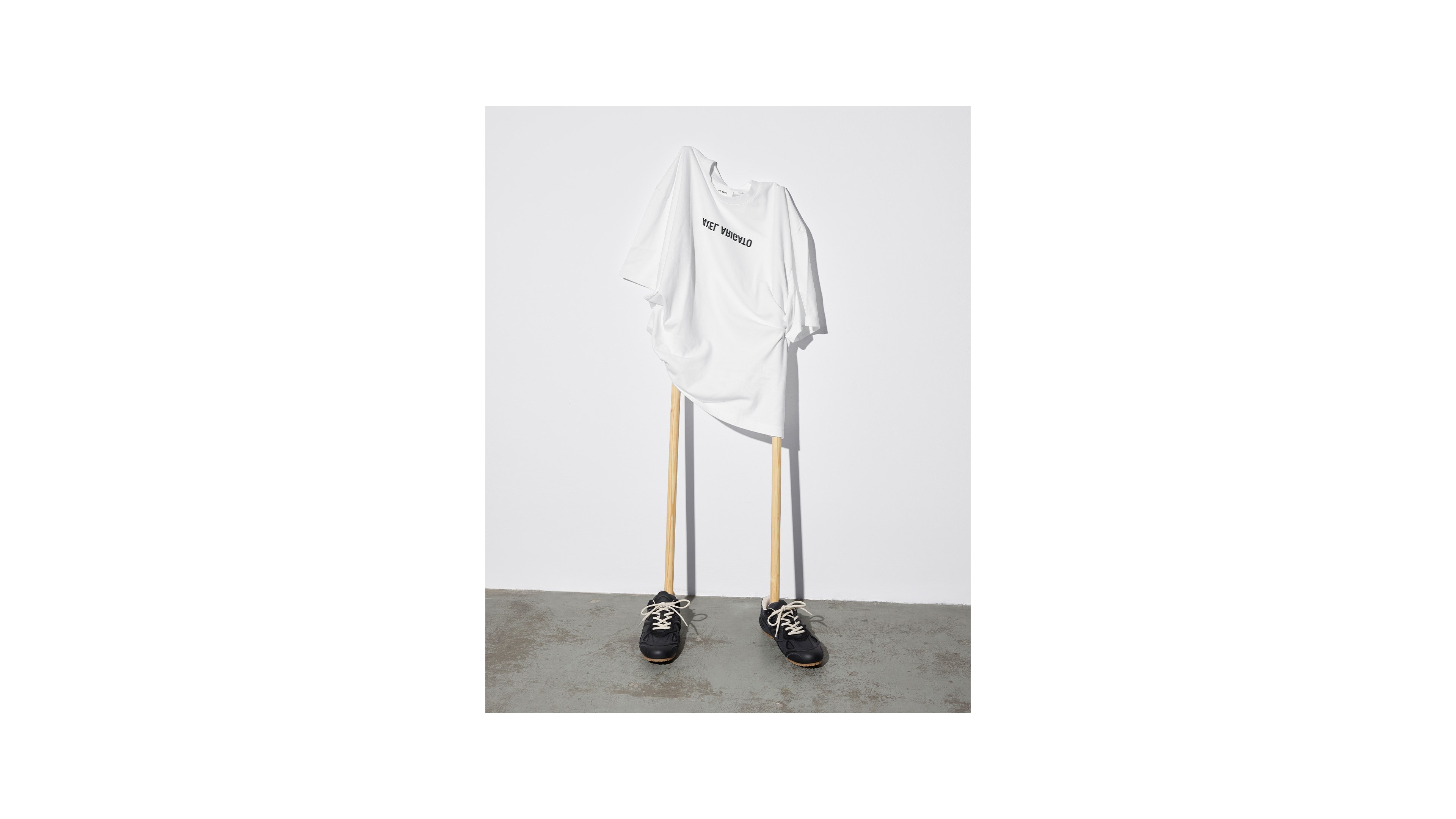 A white T-shirt on wooden poles with black sneakers at the base, resembling a minimalist human figure against a plain background.