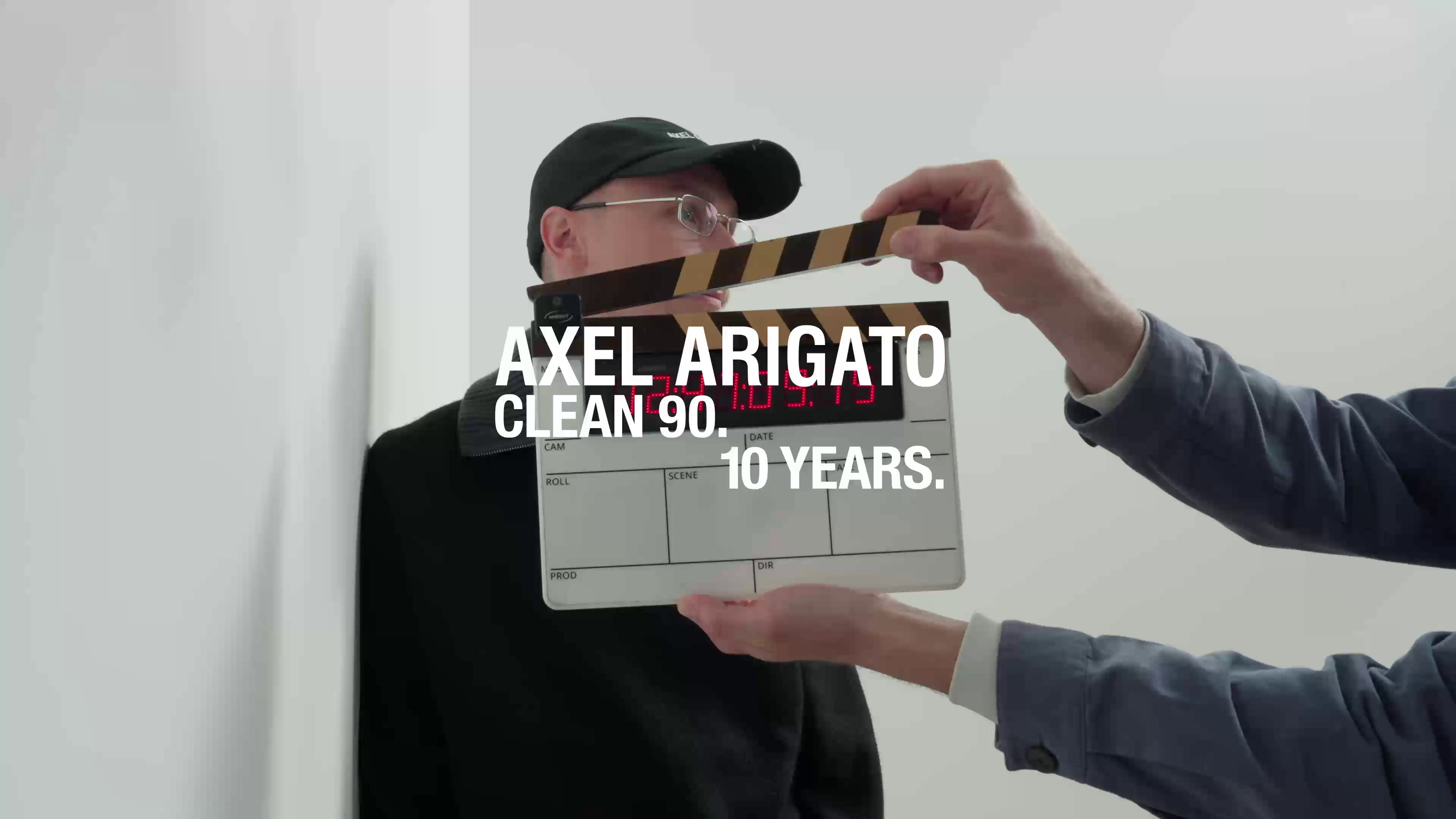 Person holding a clapperboard near a man in a black cap and glasses. Text reads "Axel Arigato Clean 90. 10 Years."