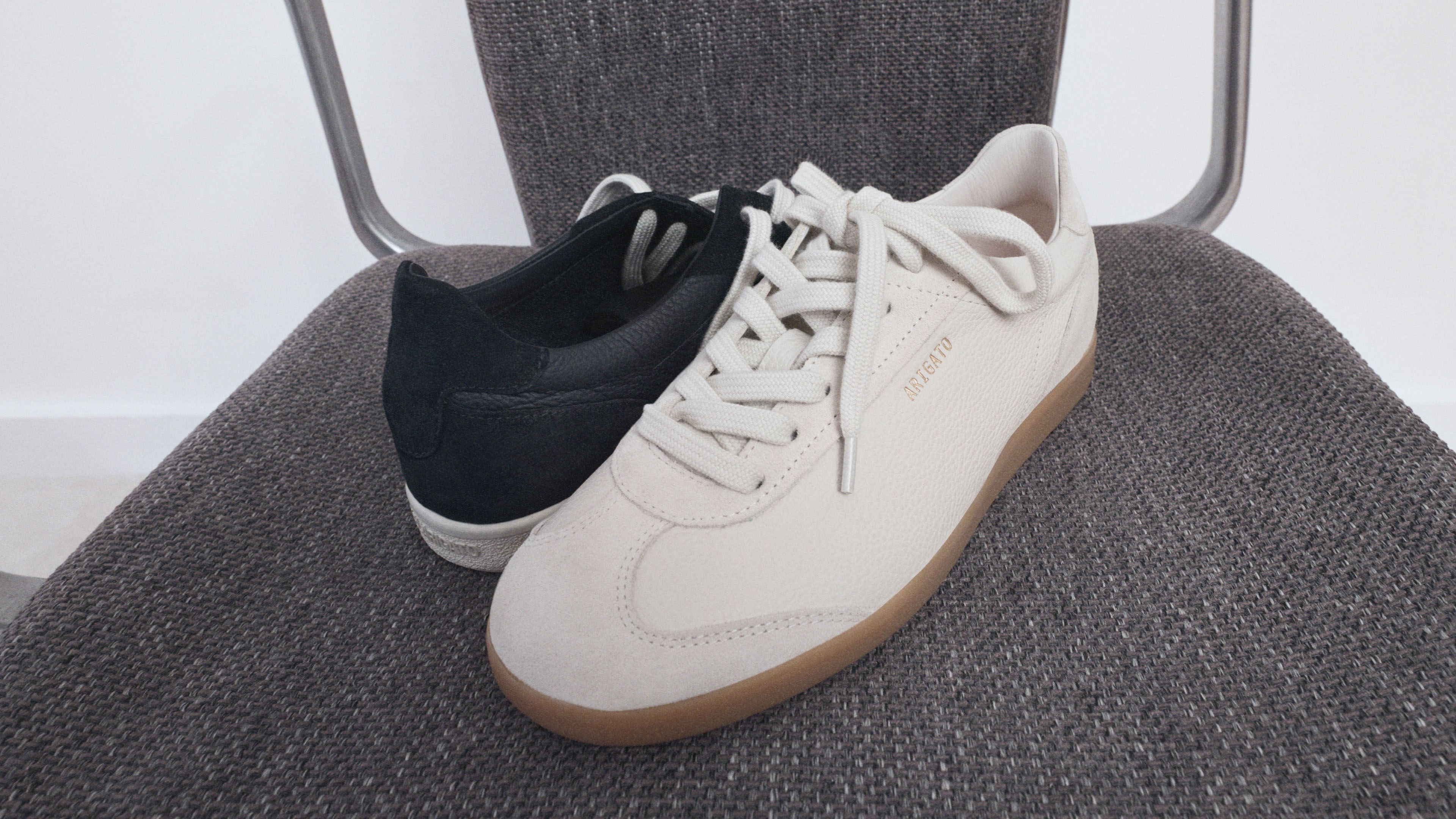 A pair of sneakers on a textured gray chair, one black and one beige, with gum soles and visible laces.