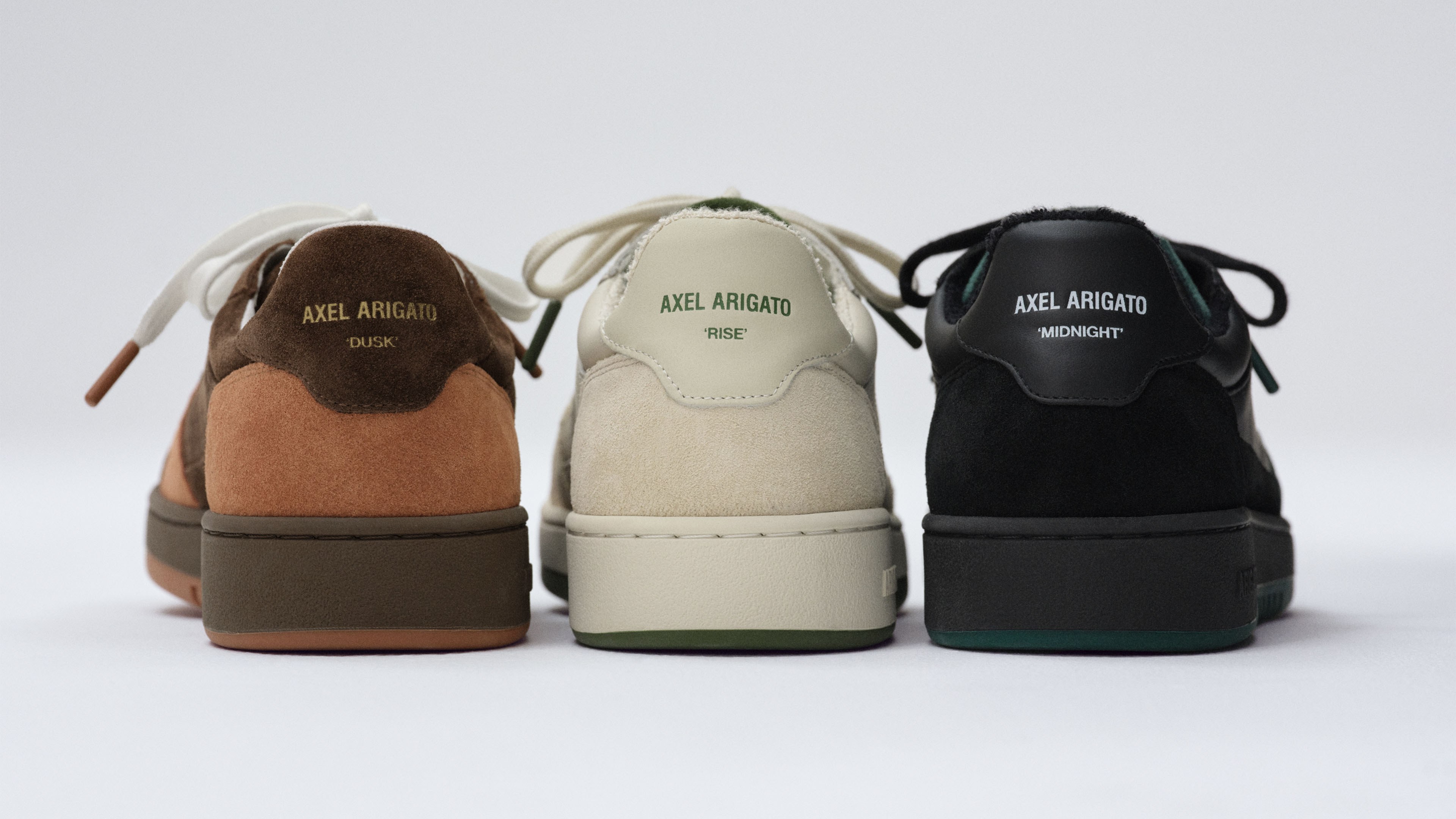 Three pairs of Axel Arigato sneakers in brown, cream, and black, viewed from the back, each with a different color sole and labeled heel.