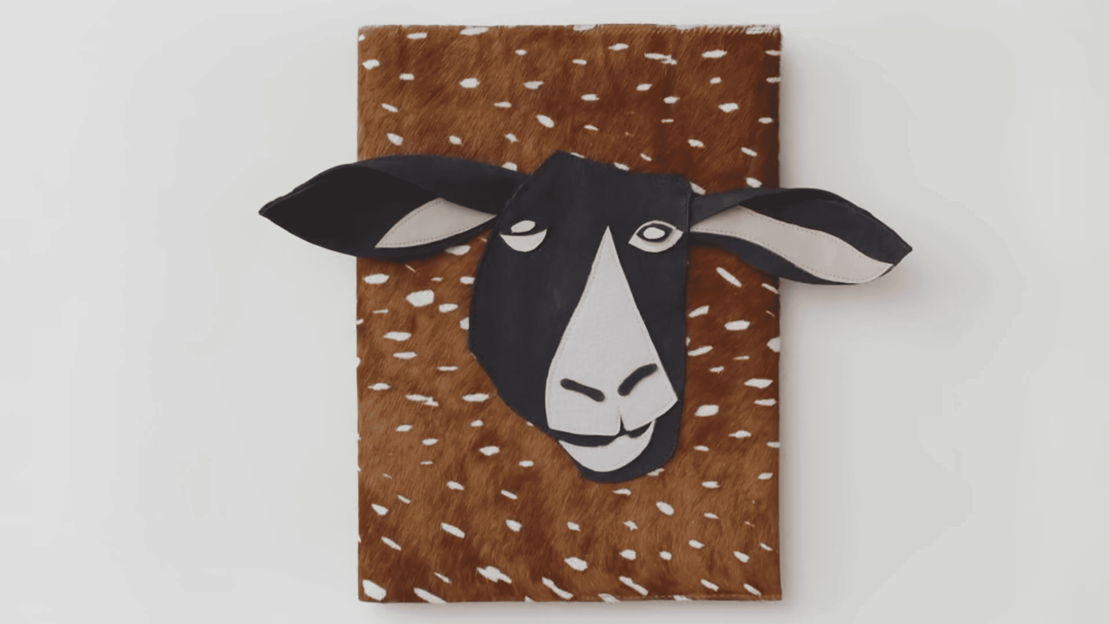A flat, textured artwork featuring a stylized goat head with black and white face and large ears, set against a brown and white spotted background.