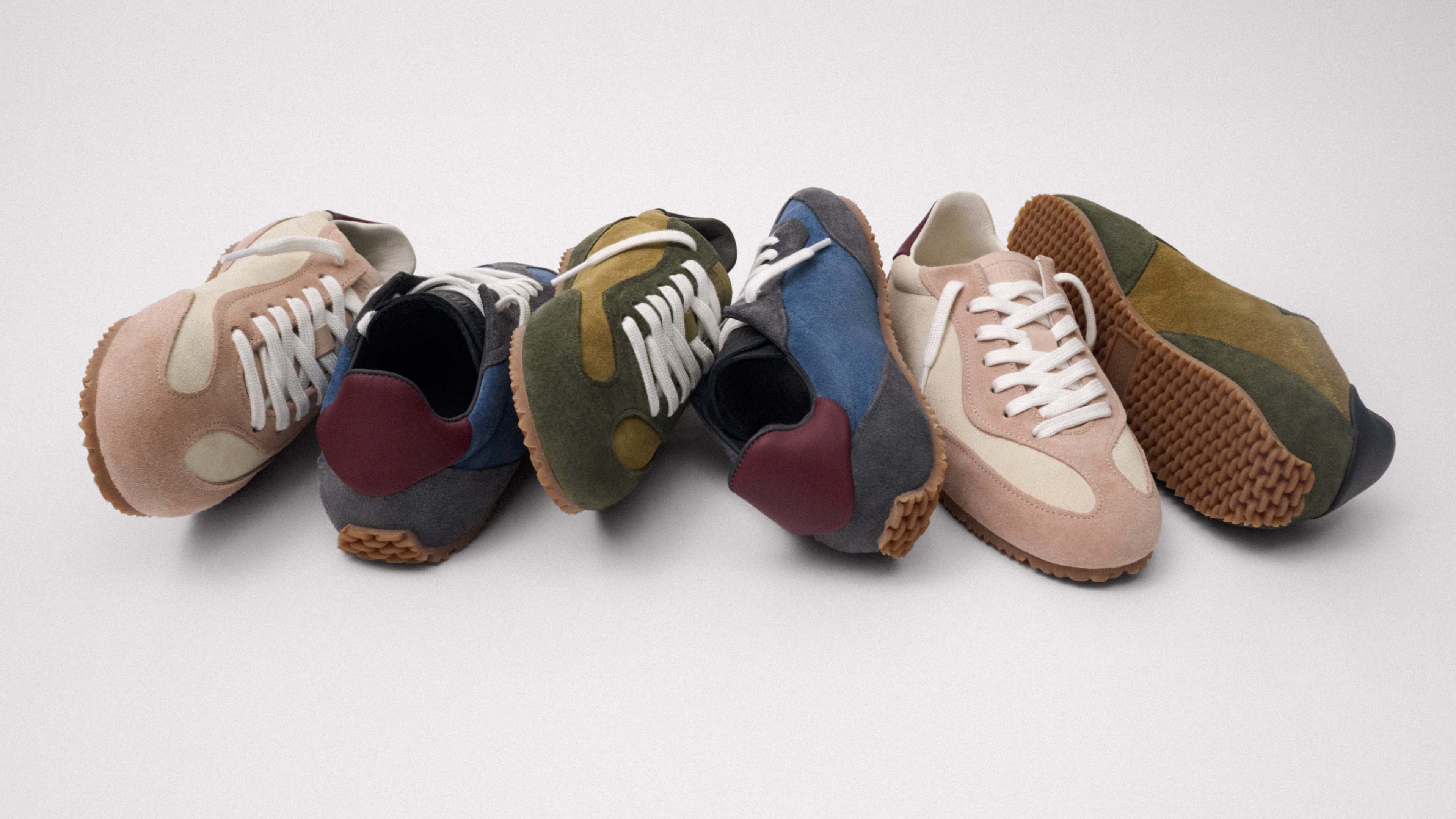 A variety of colorful suede sneakers with rubber soles, laces facing forward, arranged haphazardly on a plain gray background.