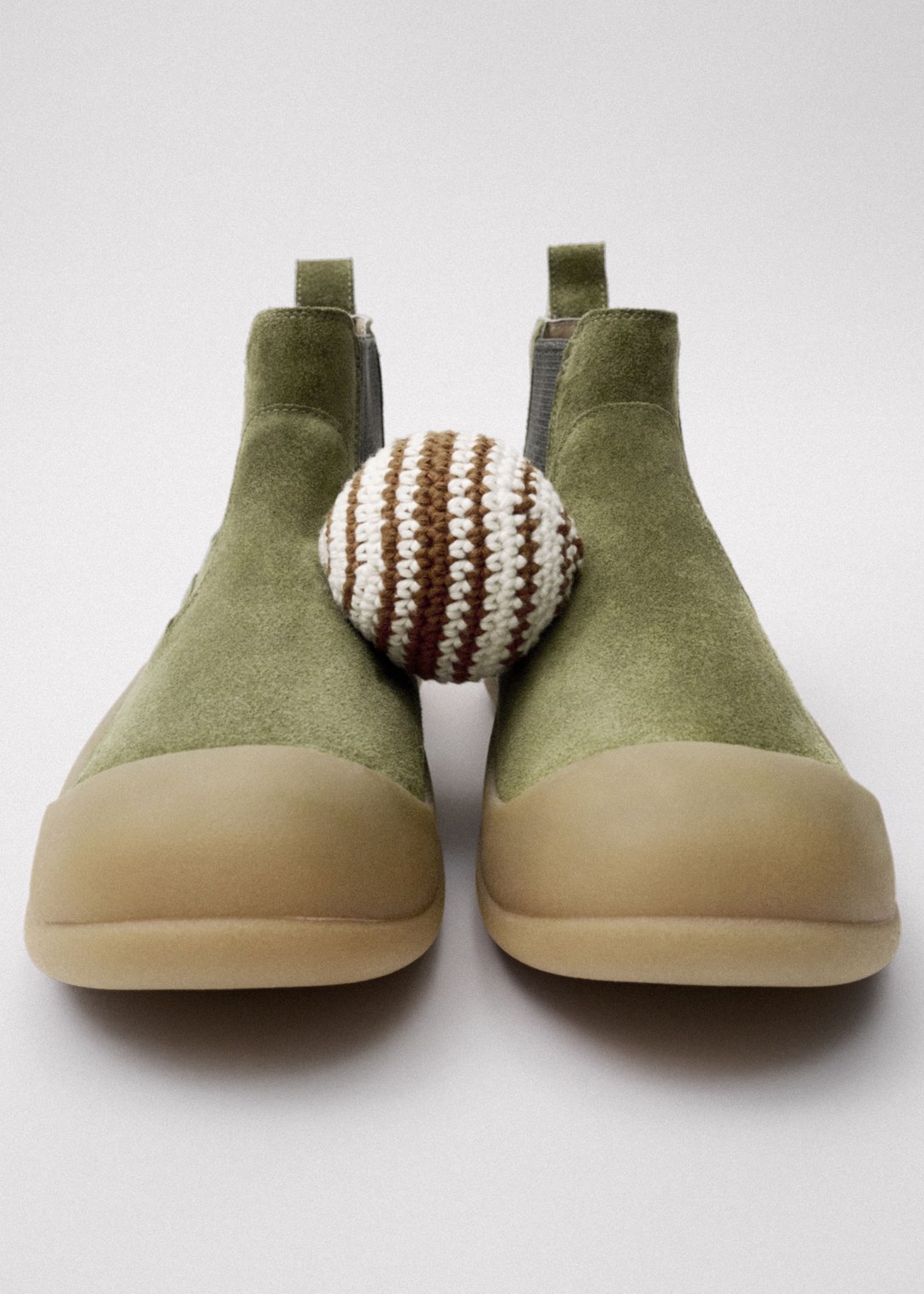 Green suede ankle boots with beige rubber soles, paired with a crocheted brown and white striped ball, placed on a neutral background.