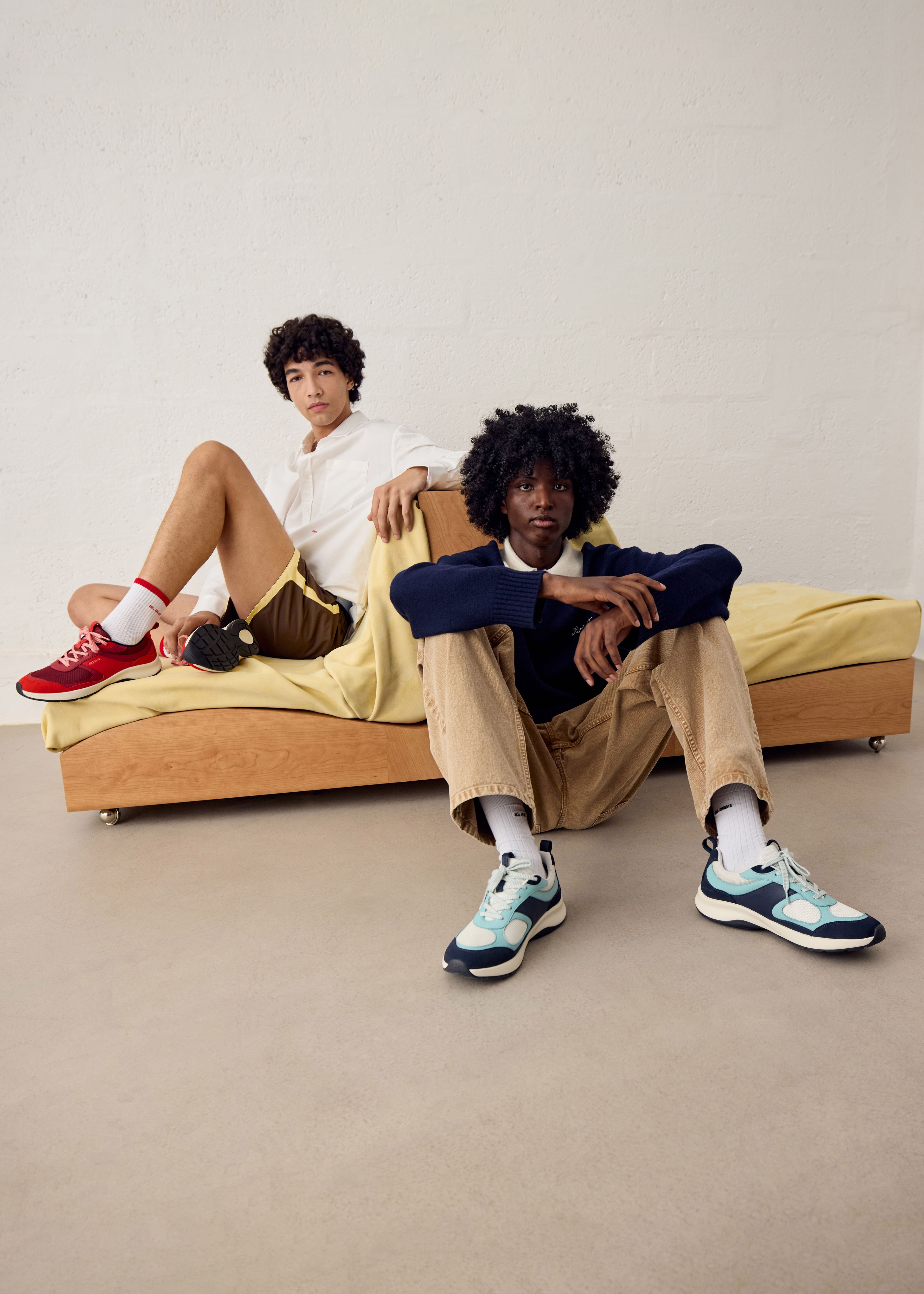 Two people posing indoors; one seated on a bed wearing a white shirt and red sneakers, the other on the floor in a sweater and blue sneakers.