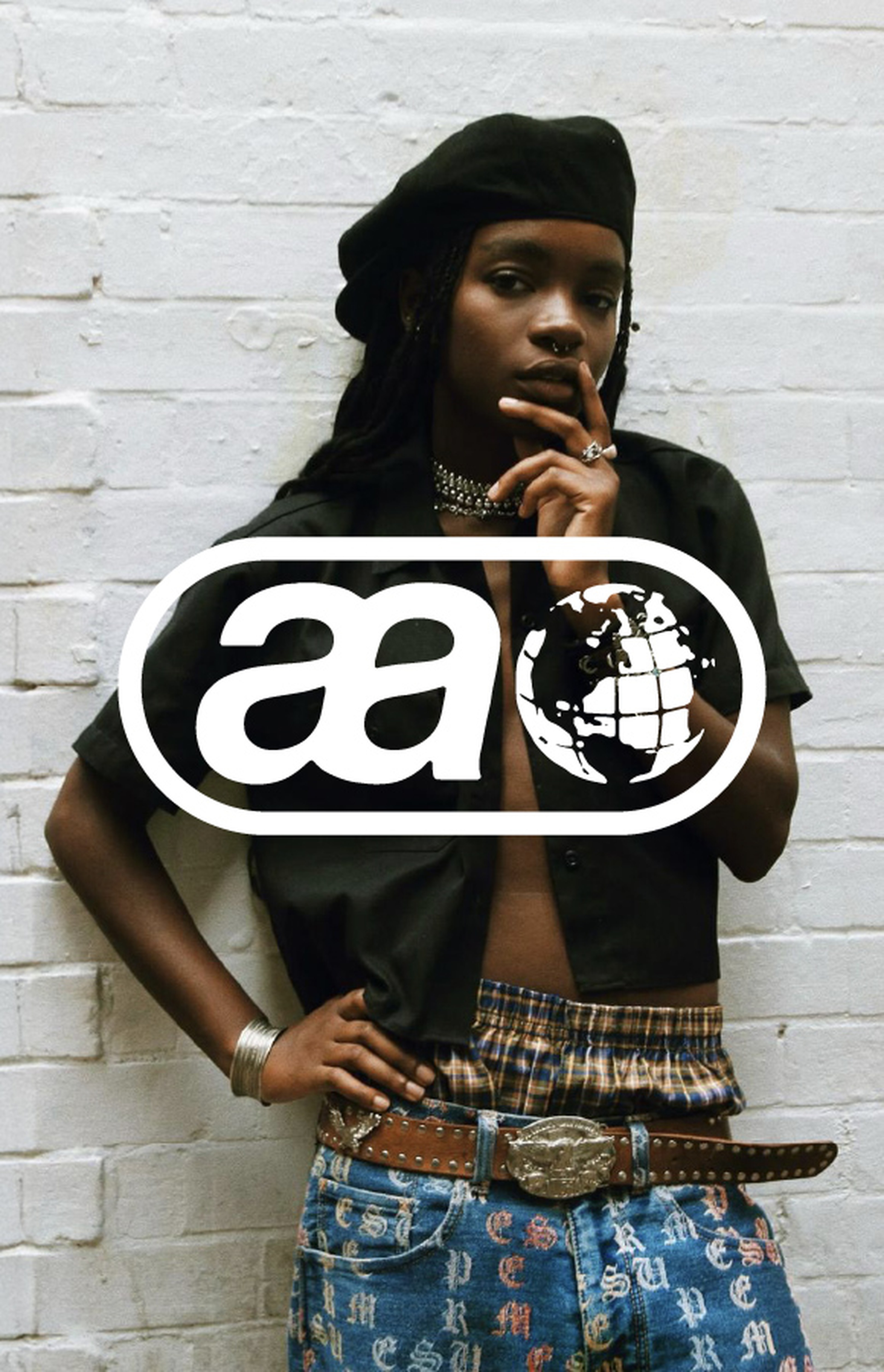 Person in stylish clothing poses against a white brick wall, wearing a beret, open shirt, plaid waistband, and jeans with colorful lettering.