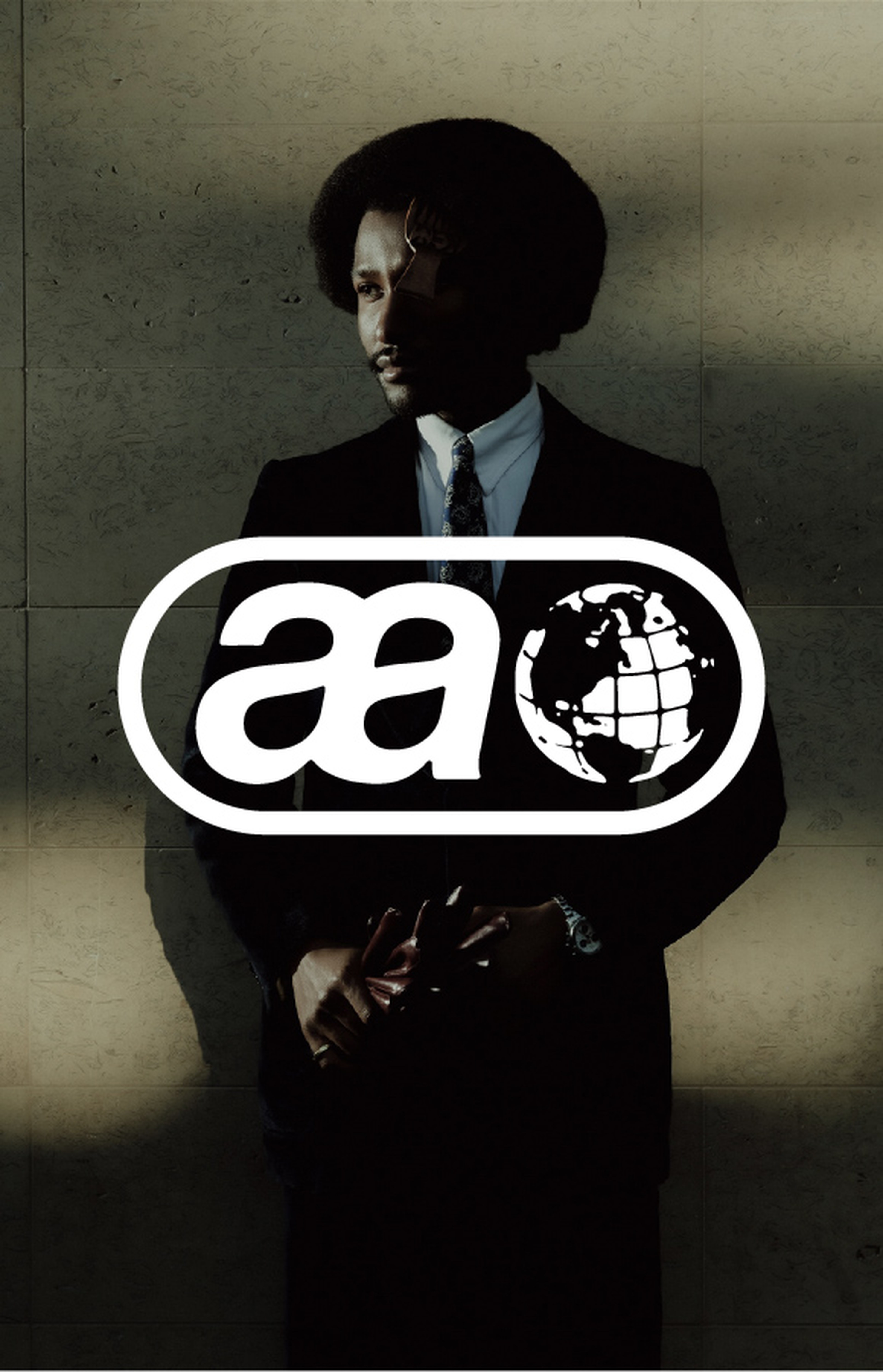 Man in a suit stands against a wall with shadows; a logo featuring "aa" and a globe is overlaid on the image.