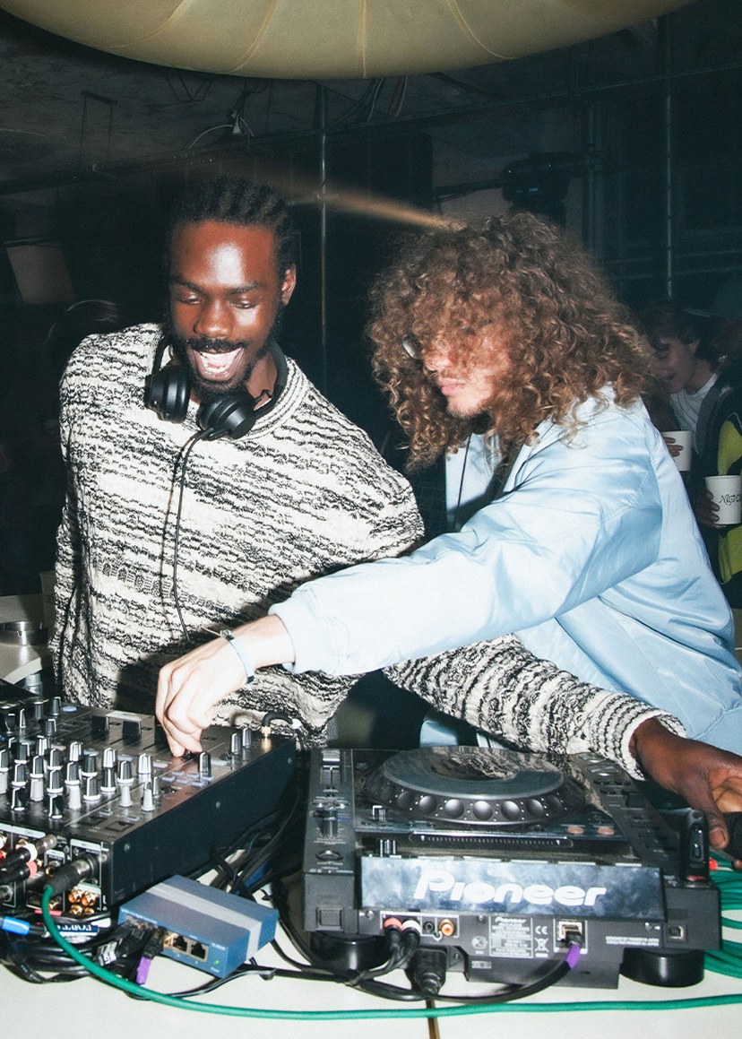 Two DJs mix music on a Pioneer turntable. One wears headphones and a striped sweater, the other a light jacket. They're in a lively club setting.