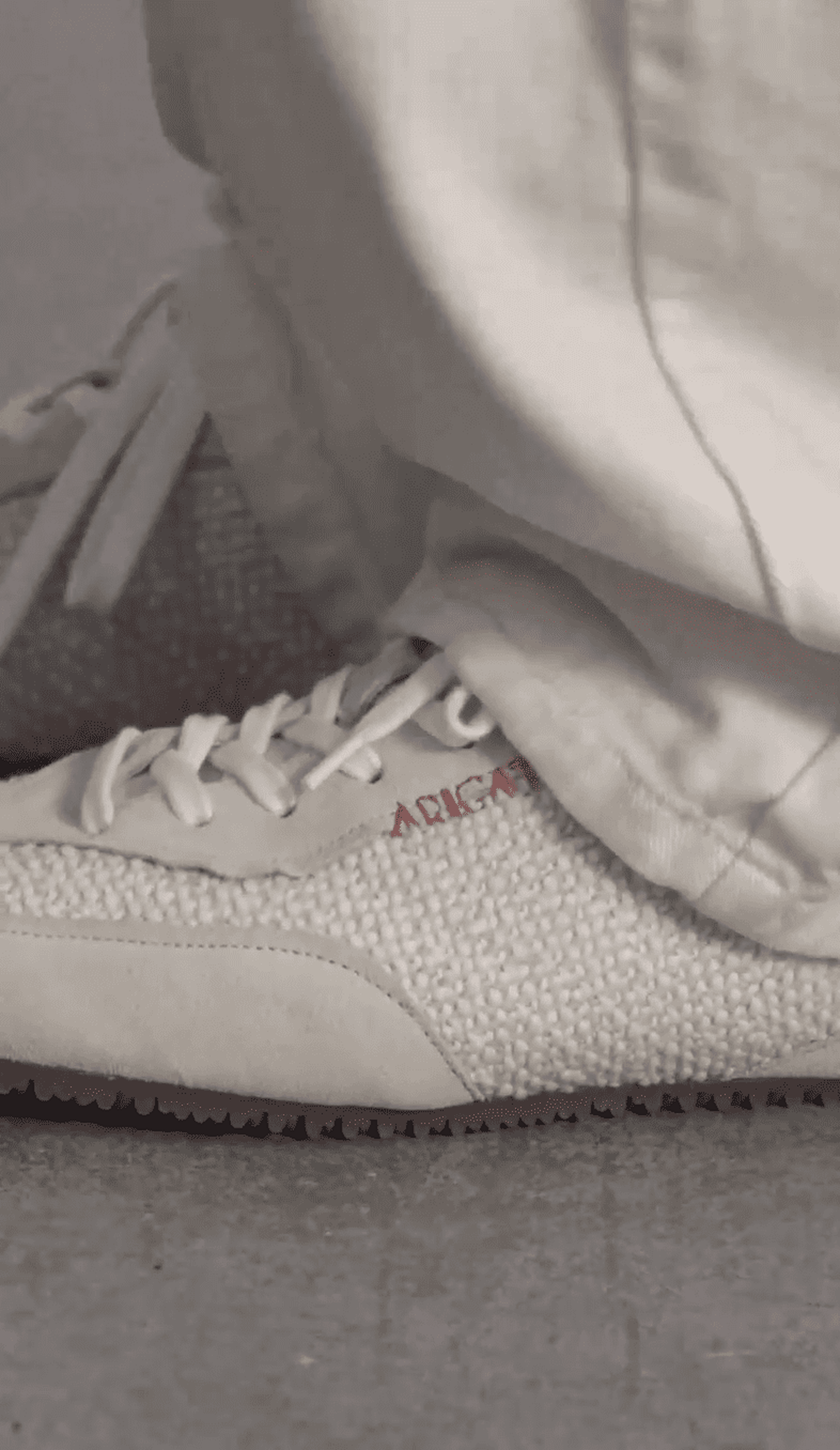 Close-up of a person wearing white textured sneakers with red lettering and loose white pants, standing on a gray surface.
