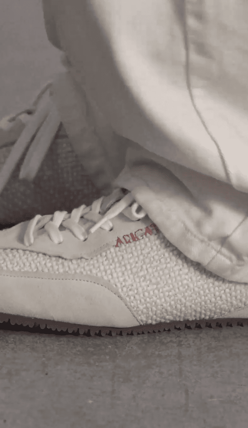 Close-up of a person wearing white textured sneakers with red lettering and loose white pants, standing on a gray surface.