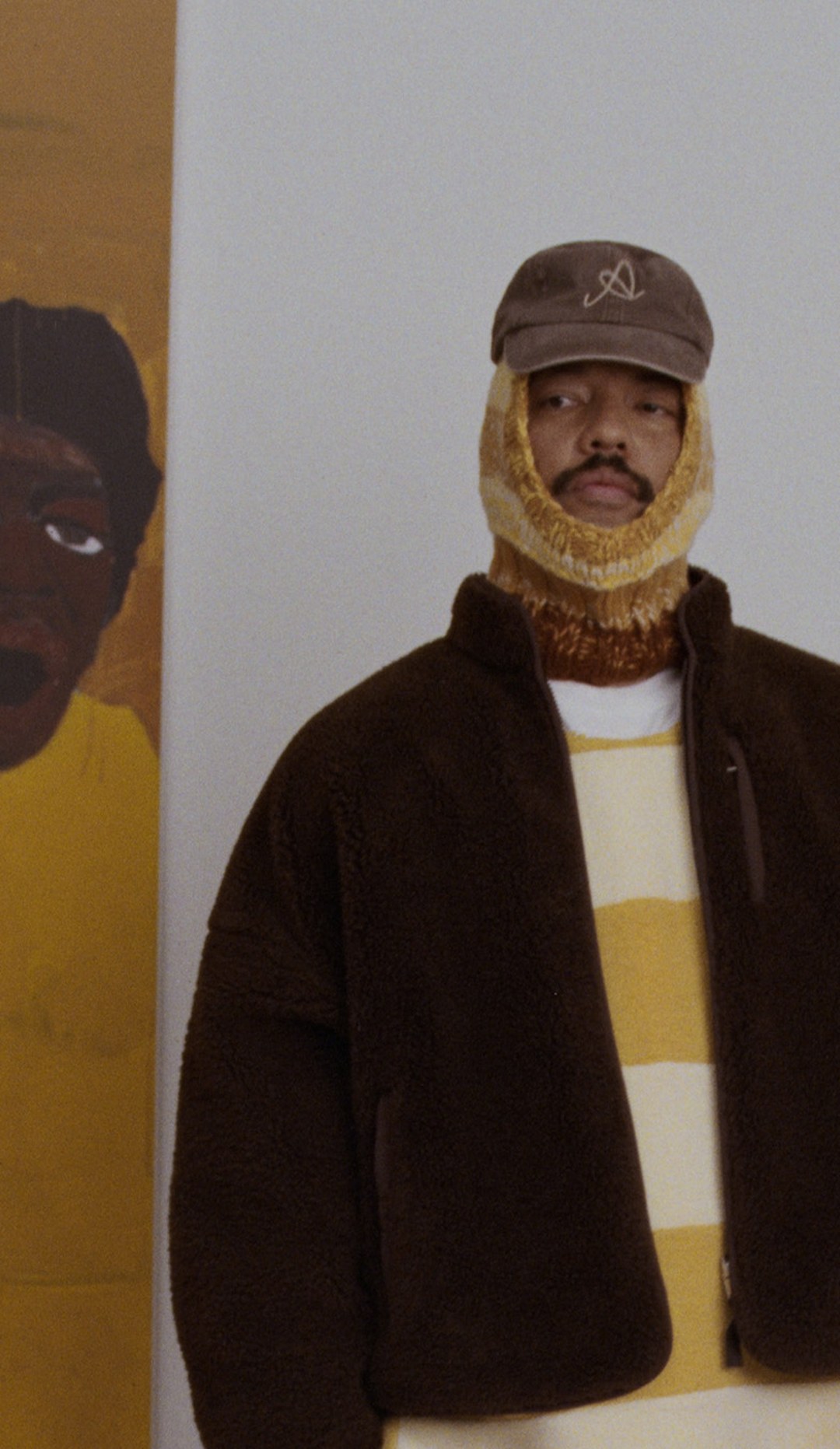 Person wearing a brown jacket, yellow and white striped shirt, and a cap with a balaclava, stands next to a painting of a face.