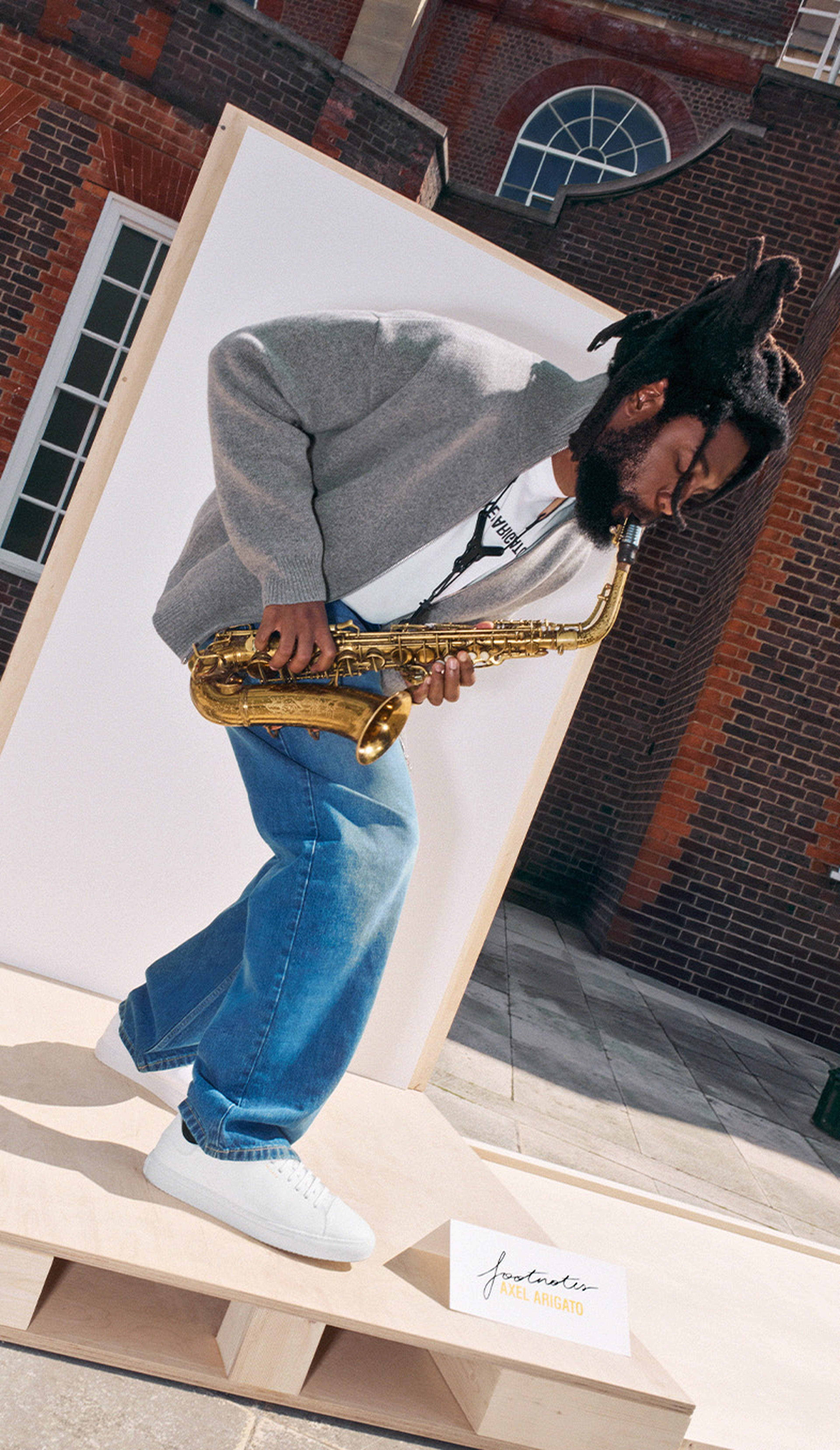 Person playing a saxophone, wearing a gray sweater, blue jeans, and white sneakers, standing on a wooden platform outdoors.
