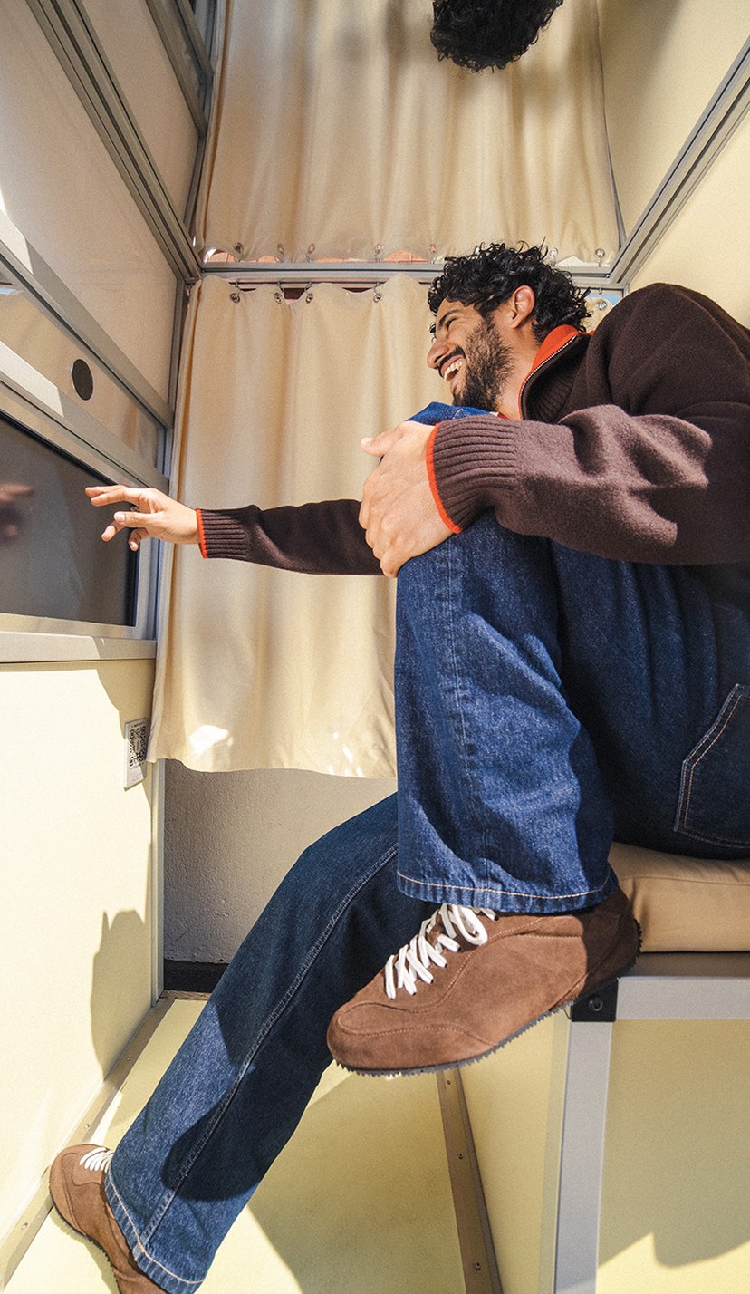 Man sitting in a small booth, smiling and looking out a window, wearing a brown sweater, blue jeans, and brown shoes.