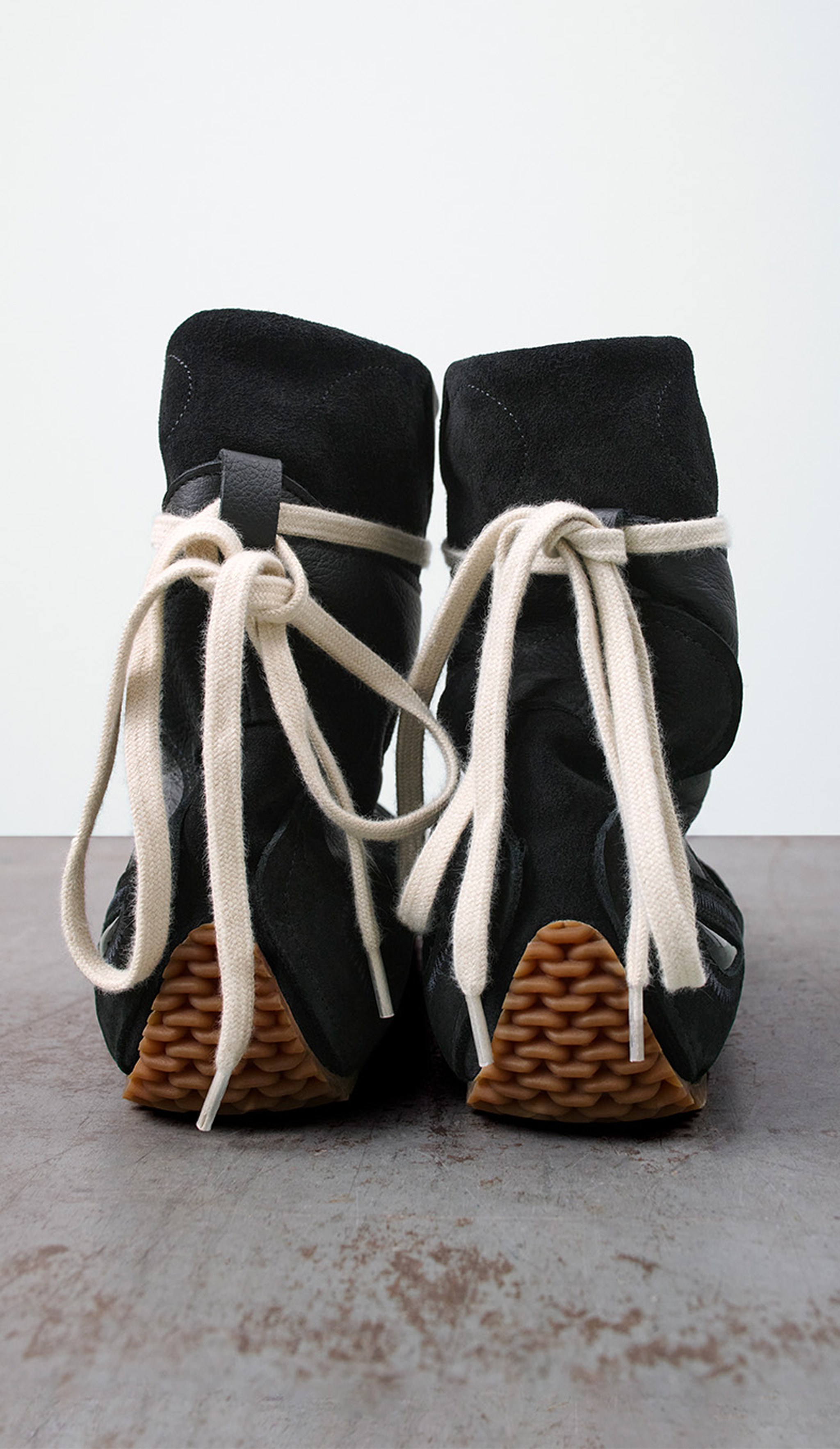 Black high-top sneakers with beige laces, featuring a textured brown sole, displayed on a gray surface.