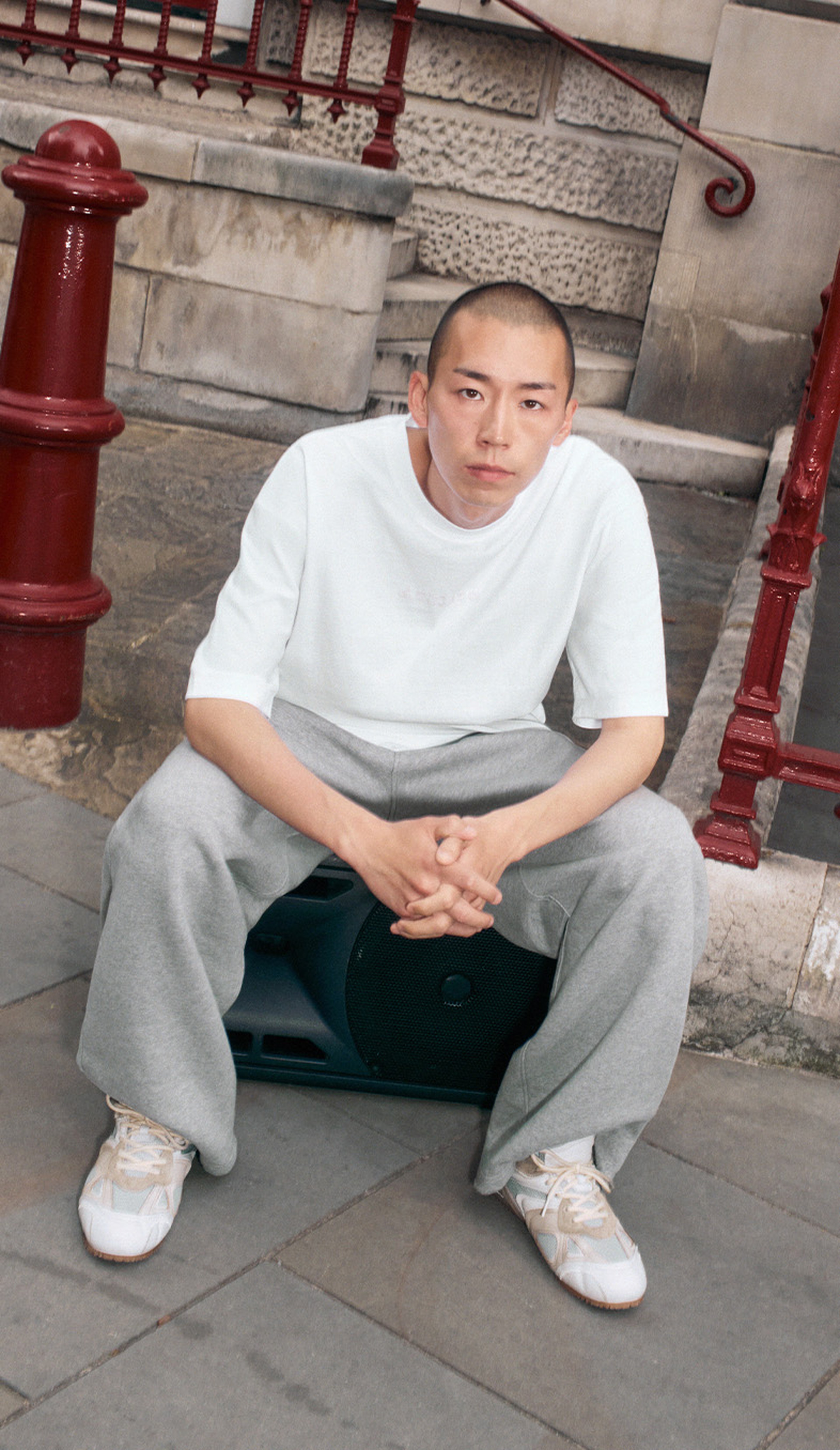 A person with a shaved head sits on a speaker, wearing a white t-shirt, grey pants, and white sneakers, in an urban setting.
