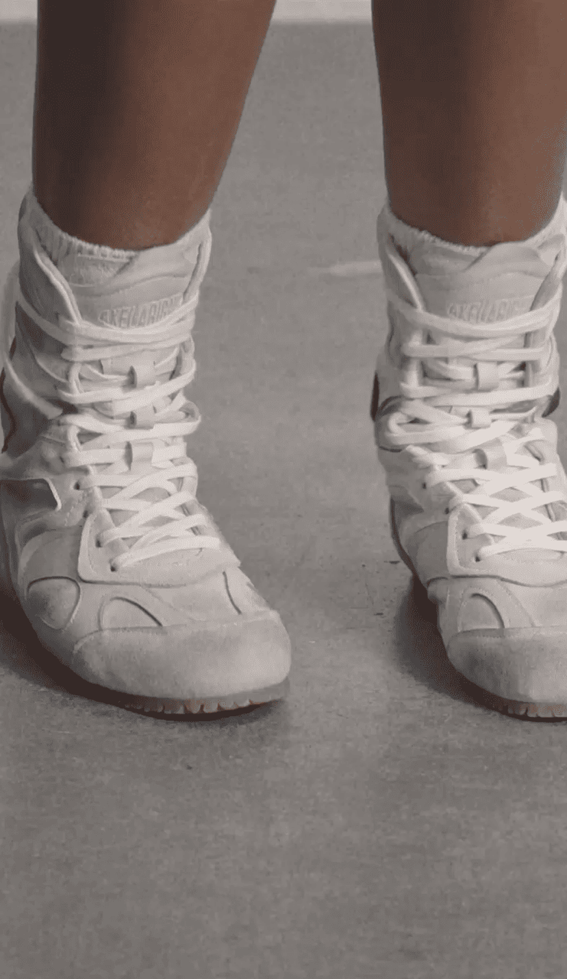 Close-up of a person wearing white boxing shoes with laces tied up, standing on a gray surface.