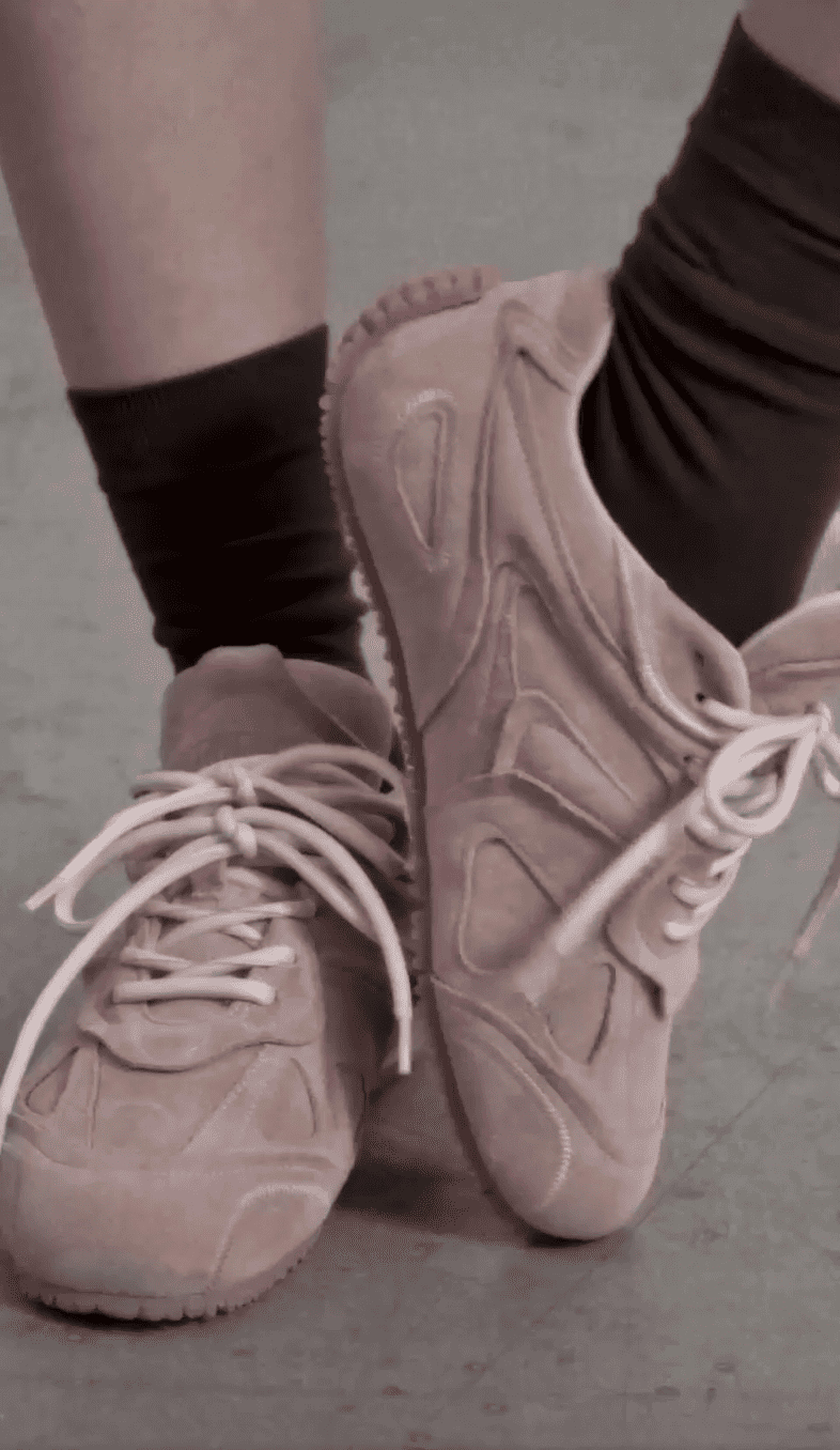 Close-up of a person wearing light pink suede sneakers with white laces, paired with dark socks, standing on a gray floor.