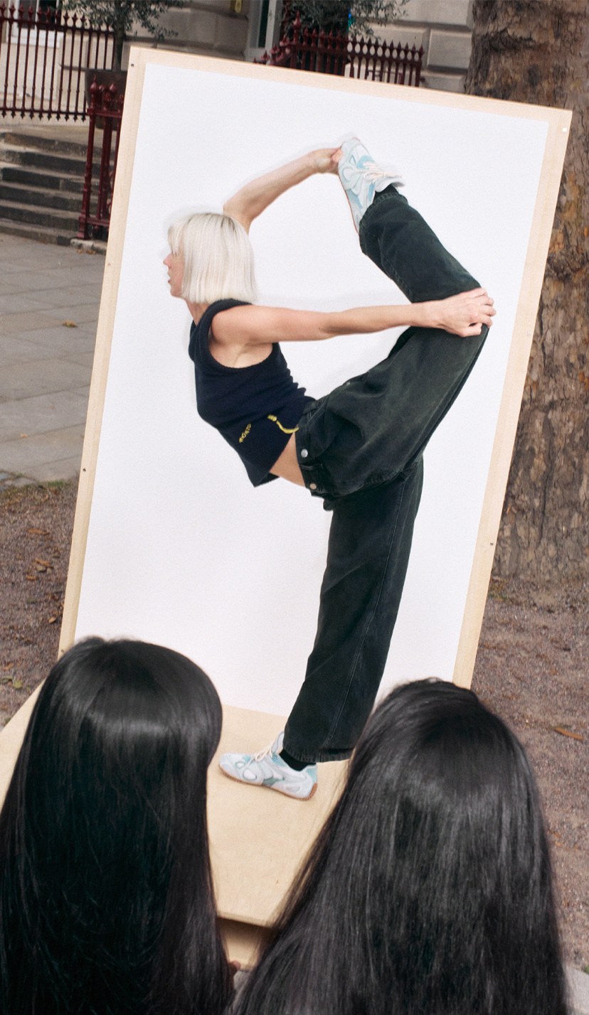 A person performs a standing split pose in front of a large mirror outdoors, observed by two people with long black hair.
