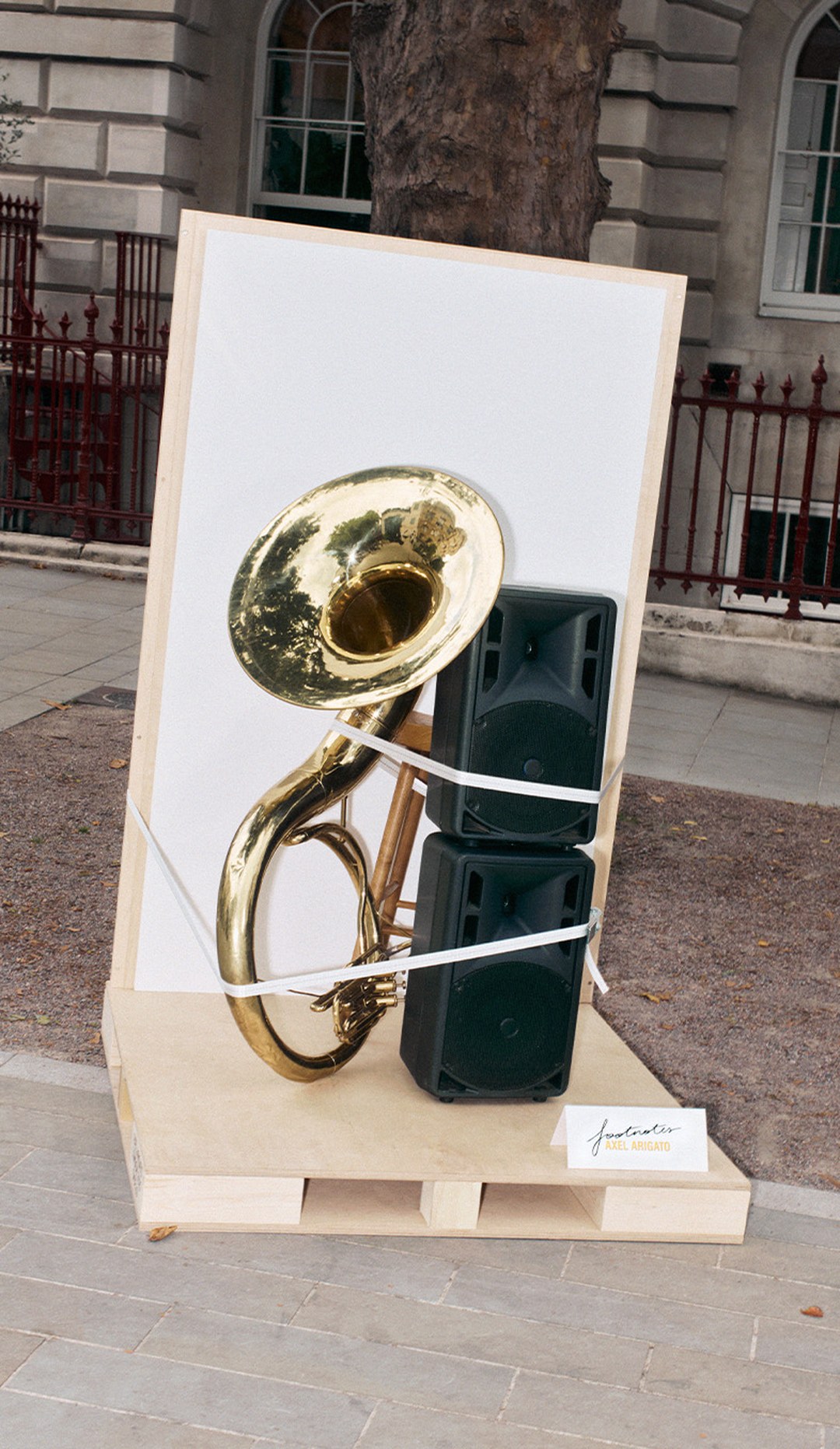 A brass tuba is strapped to a black speaker, placed upright on a wooden pallet outdoors, with a white backdrop and a small sign nearby.