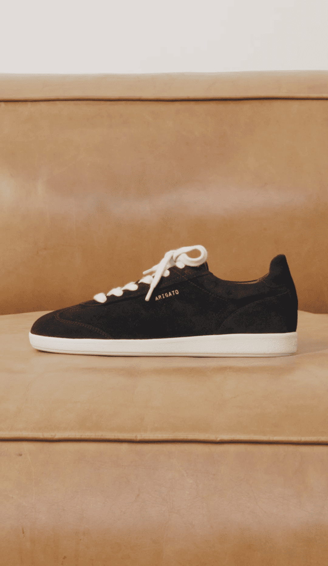 Black sneaker with white laces and sole, "Arigato" written on the side, placed on a tan leather couch.