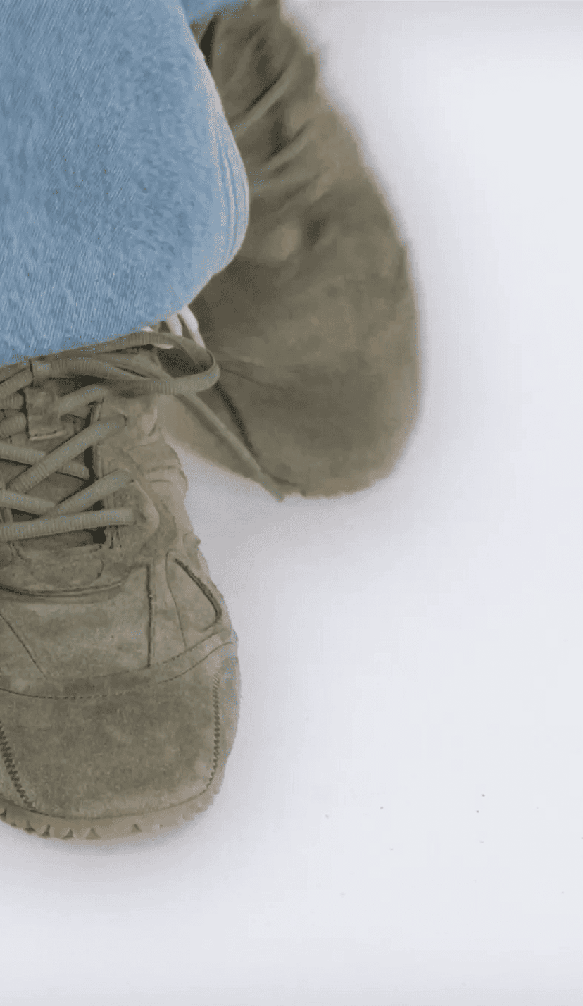 Close-up of person wearing brown suede sneakers and blue jeans, sitting with legs crossed on a white background.