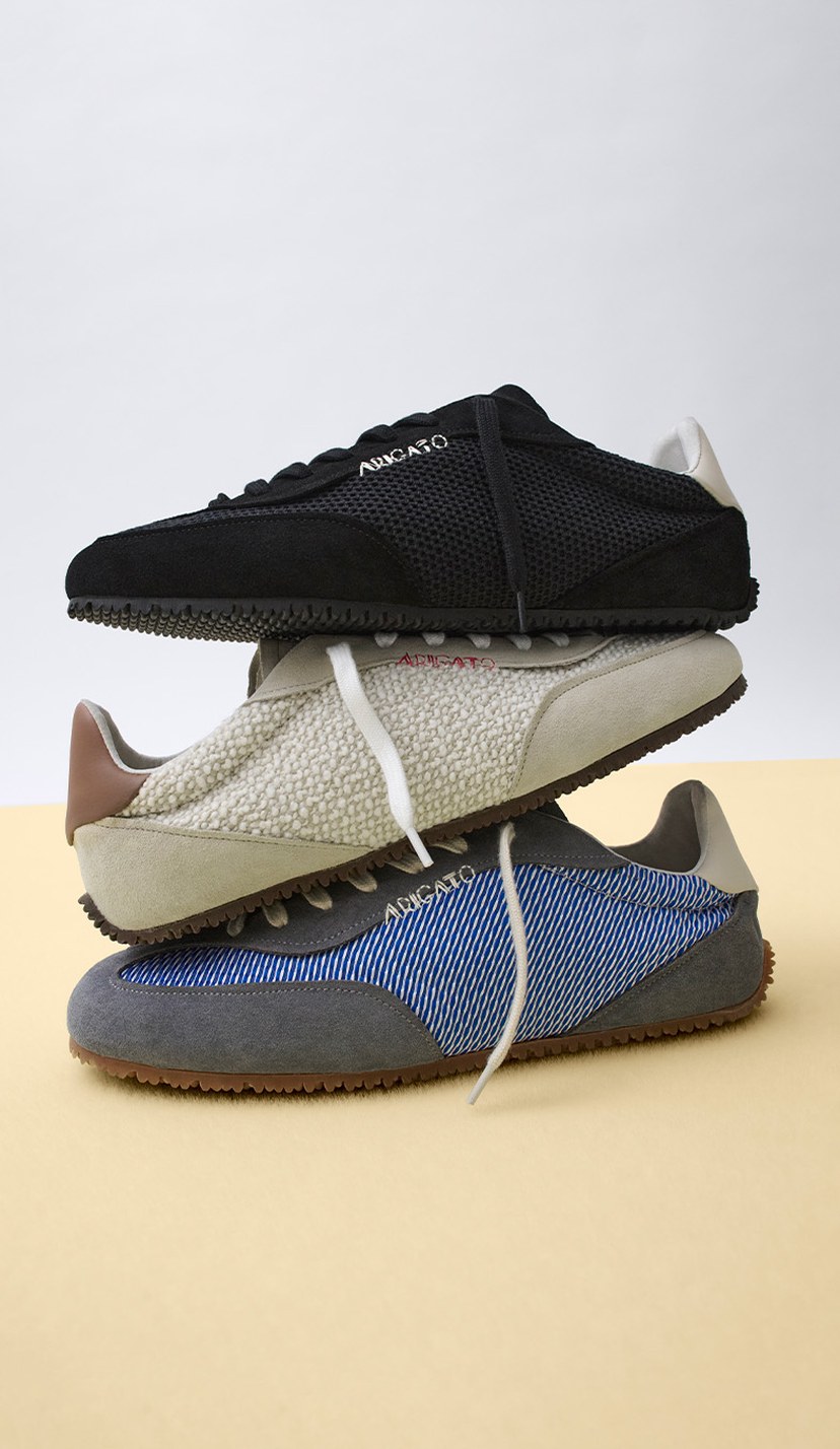 Three sneakers stacked on a yellow surface: black on top, beige in the middle, and blue with white stripes on the bottom.