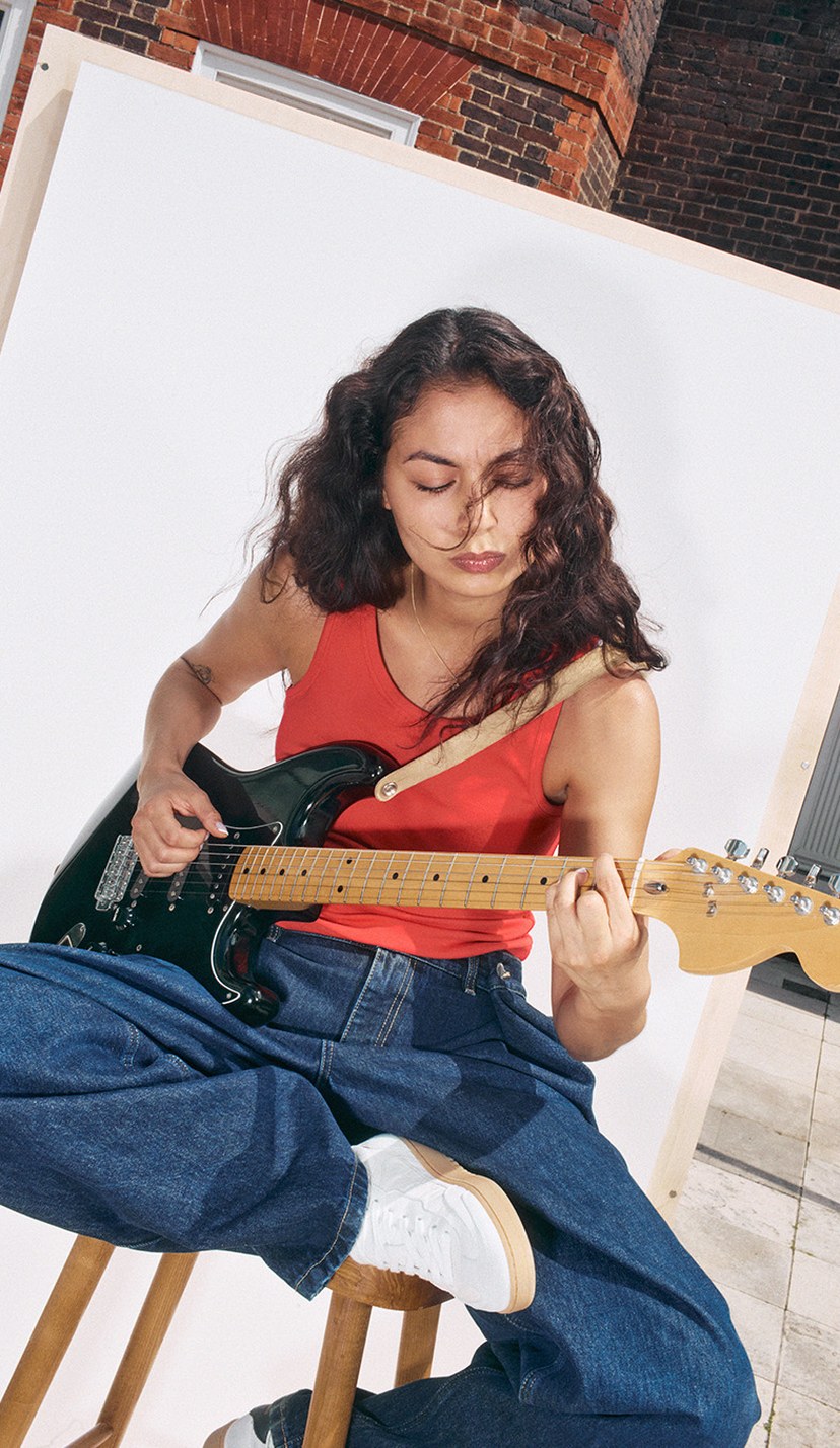 A woman with long curly hair plays an electric guitar while sitting on a stool, wearing a red tank top and jeans, in an outdoor setting.