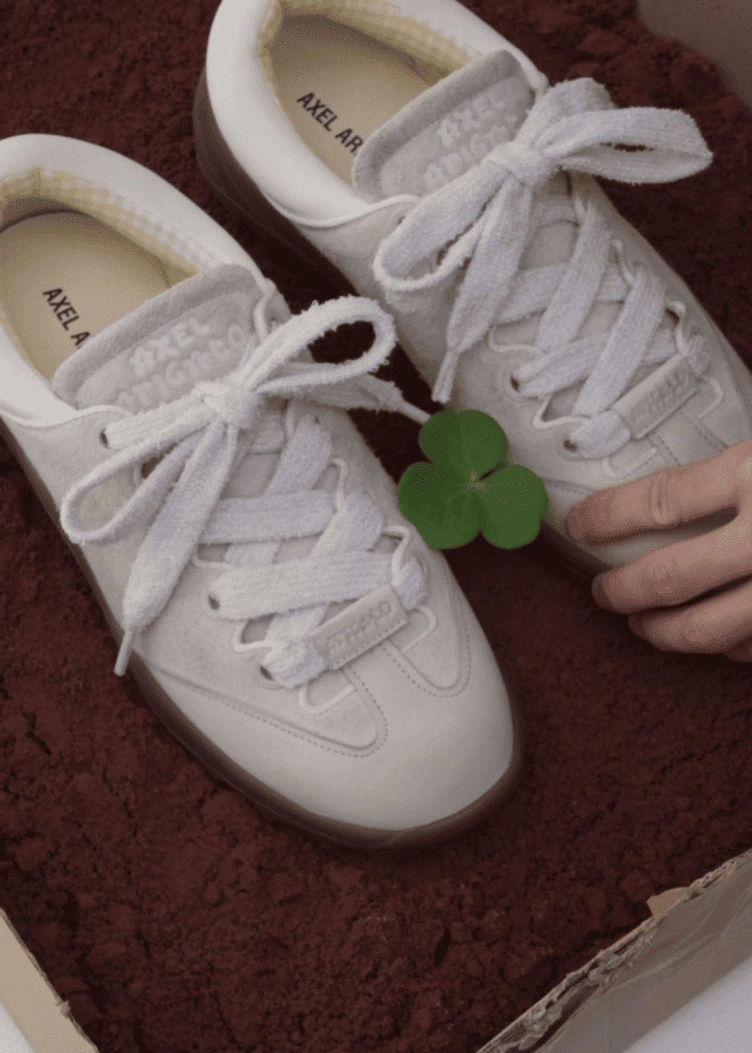 White sneakers with thick laces rest on a bed of dark brown soil. A small green four-leaf clover lies atop the shoes.
